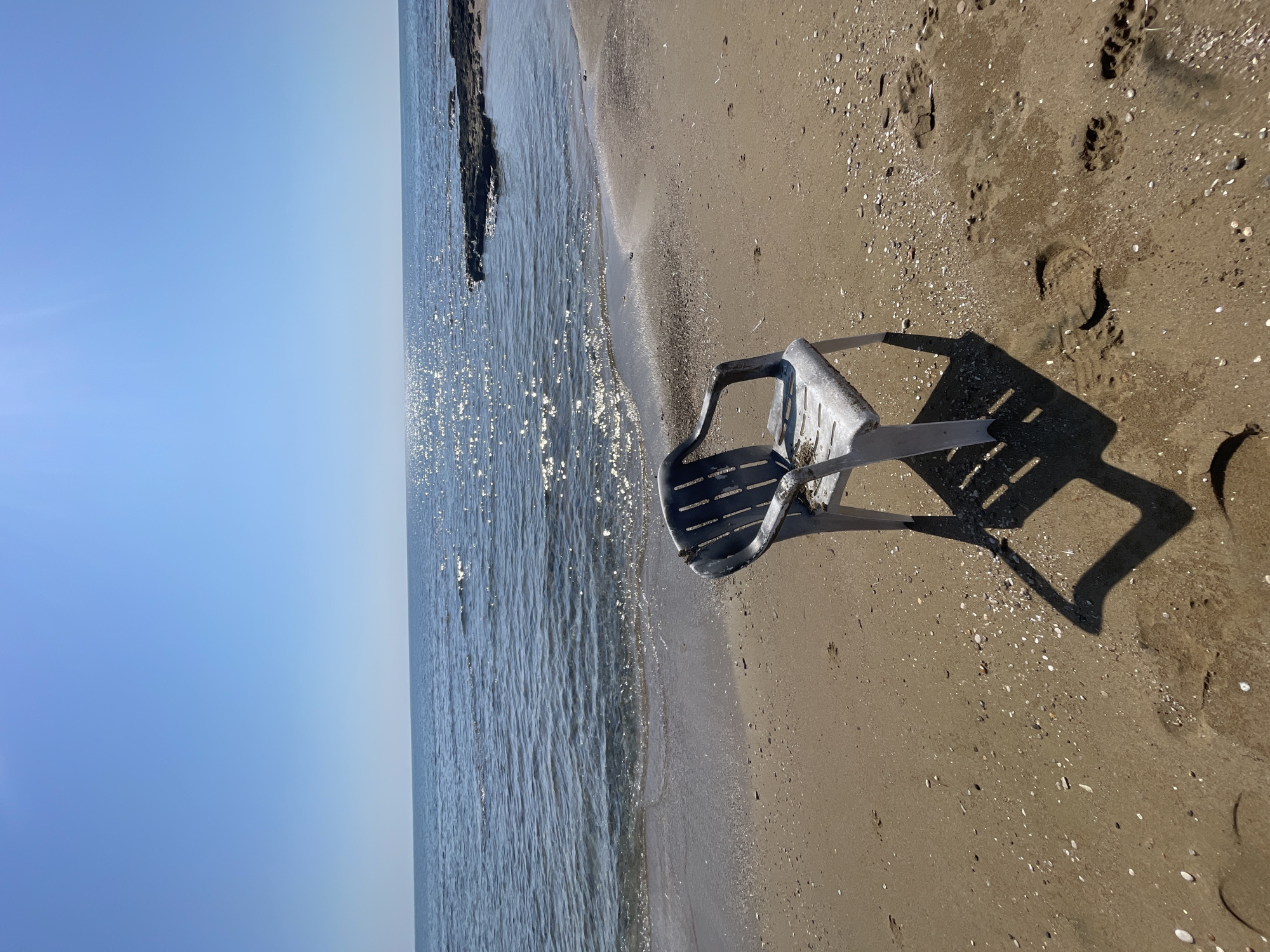 The image depicts a serene beach scene on a clear, sunny day. In the foreground, a weathered white and gray plastic chair sits on the sandy shore. The chair casts a distinct shadow on the sand. The beach is sparsely scattered with small seashells and pebbles. Footprints are visible in the sand, suggesting recent human presence. The gentle waves of the blue sea lap at the shore, with sunlight shimmering on the water's surface. In the background, a rocky outcropping extends into the sea. The sky is a clear, unbroken blue. The scene is likely located in Agios Sergios, Northern Cyprus, a region known for its picturesque coastline.