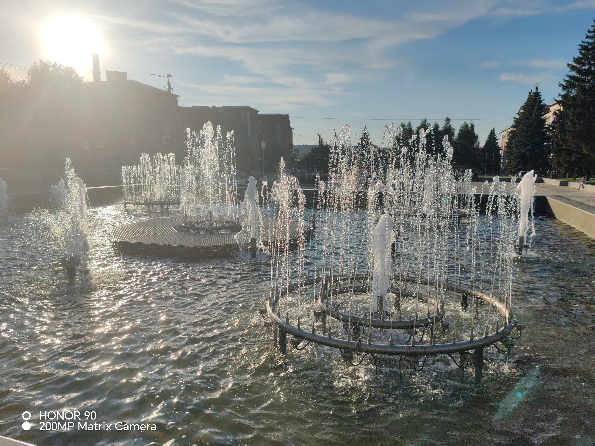 A bright, sunny day is captured at a fountain in an urban setting. The sun, positioned in the upper left quadrant of the image, is partially obscured by a bright lens flare. Numerous water jets from an intricate fountain system erupt from circular metal structures in the foreground, creating a shimmering, translucent effect as they catch the sunlight. The water surface is rippled, reflecting the bright sky and the spray from the fountain. In the background, several buildings are visible, with a tall industrial chimney to the left. Trees, including several tall evergreen trees, line the right side of the scene, bordering a paved walkway. The sky is blue with scattered white clouds. A portion of a tiled walkway or plaza is visible to the right of the fountain. A person is faintly visible walking on the walkway in the distance. The lower portion of the image contains a watermark that reads "HONOR 90 200MP Matrix Camera."