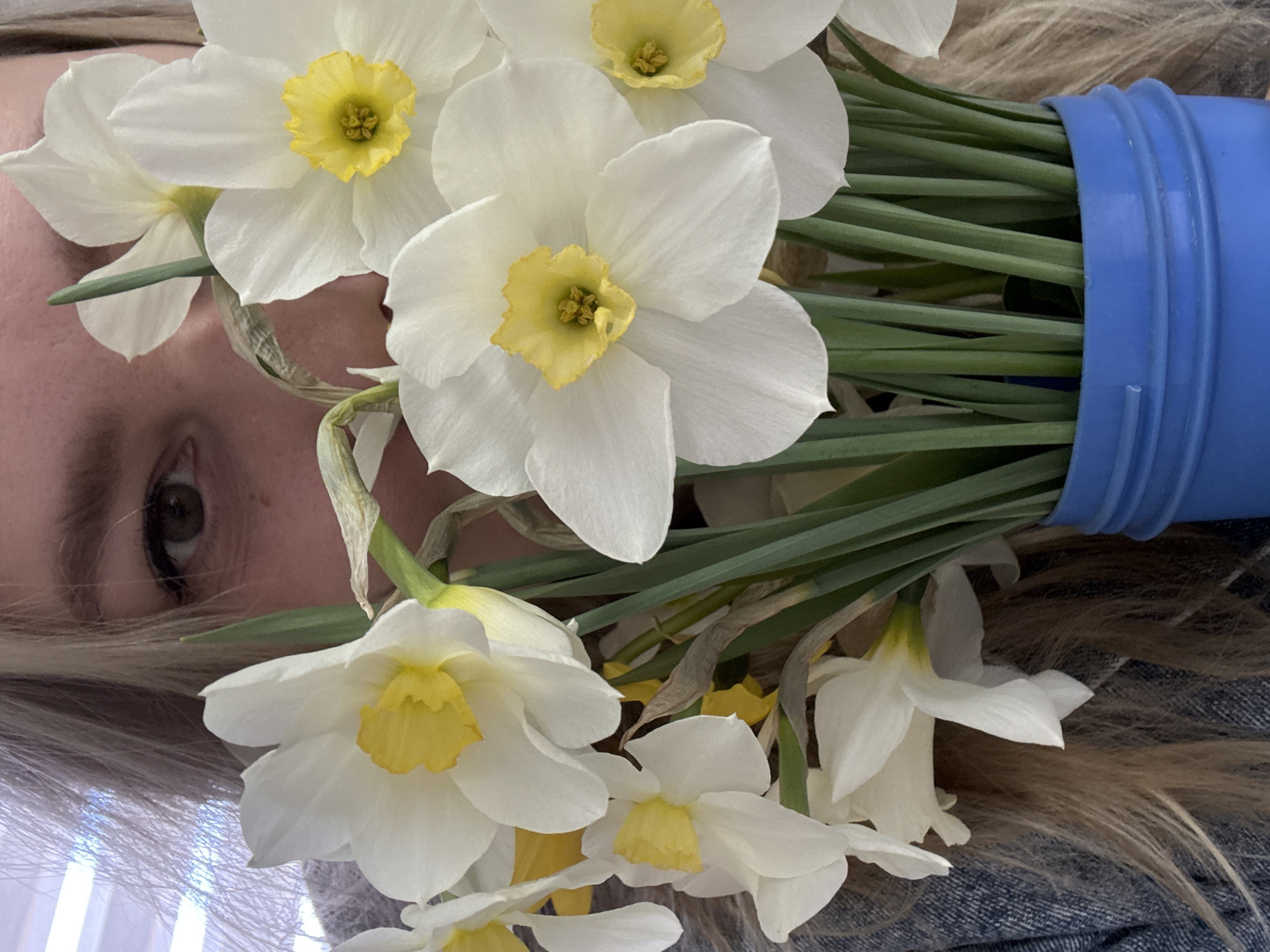 A person's eye, blonde hair, and a blue container filled with white and yellow daffodils are visible. The scene is indoors with a light source coming from the left. The person appears to be lying down or positioned such that the daffodils obscure much of their face, with the eye visible in the upper left quadrant of the image. The lower half of the image is filled with white and yellow daffodils. The blue container is in the upper right quadrant. The image's location is unknown and cannot be determined from the available information.