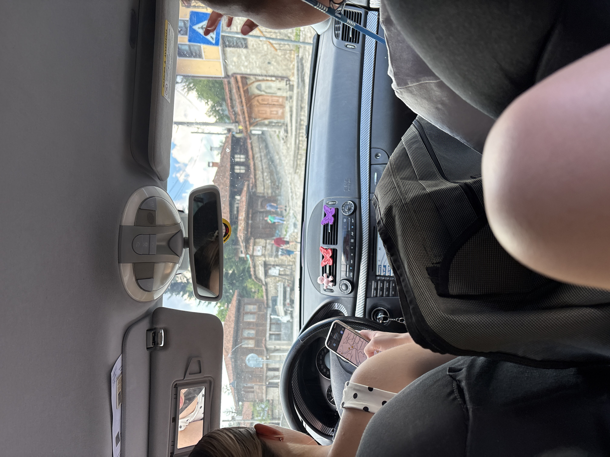 This media file shows the interior of a car, viewed from the passenger's perspective, as it drives through the historic town of Koprivshtitsa, Bulgaria. The car's interior is moderately lit, suggesting it is daytime. The sun visor on the driver's side is flipped down, and a person's hand is visible near it, holding a blue object. The dashboard features a car's stereo system and air vents, with a purple butterfly-shaped air freshener attached. A smartphone is mounted on the dashboard, displaying a map, indicating the car is navigating.
The view through the windshield reveals a street scene with traditional Bulgarian architecture. Buildings with distinctive red tile roofs and stone facades are visible, characteristic of Koprivshtitsa. The presence of a blue triangular road sign with a white arrow suggests navigation is in progress. The reflection in the rearview mirror shows a person, possibly the driver, with light-colored hair and wearing a white top with black polka dots. Another person's leg is visible in the foreground, wearing dark pants. The overall impression is of a casual journey through a picturesque historical location.