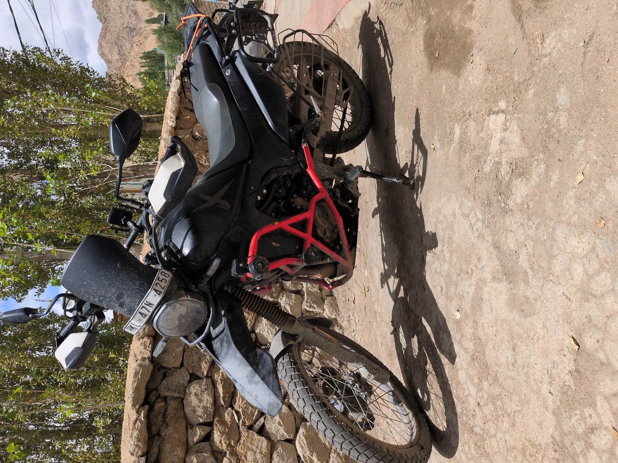 A black Royal Enfield Himalayan motorcycle is parked outdoors on a dirt and paved surface in Leh, India. The motorcycle has a tall windshield, handguards, a red crash cage, and a luggage rack at the rear. The front wheel is a spoked wheel with a knobby tire, and the rear wheel is also a spoked wheel. A license plate reading "KL 47N 4258" is visible on the front of the motorcycle. A stone wall is behind the motorcycle, and trees with green foliage are in the background. Mountains and a few buildings are visible in the distance under a cloudy sky. The scene appears to be a typical roadside setting in the mountainous region of Leh.