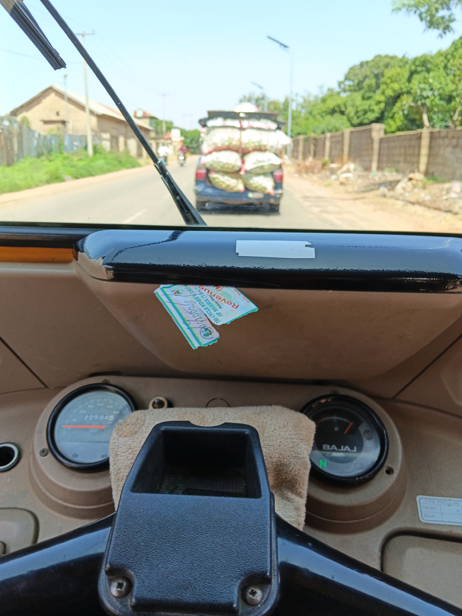 The image is taken from the inside of a vehicle, looking forward through the windshield. The steering wheel and dashboard are visible in the foreground. The dashboard has two gauges, one showing "225942" and the other labeled "BAJAJ". A small piece of torn paper with text in green and white is stuck to the dashboard. A beige cloth is draped over the top of the gauges.
Outside the vehicle, a road is visible stretching into the distance. A dark blue vehicle is in front, heavily laden with large, white sacks, possibly produce. Trees and buildings line the sides of the road. The sky is clear and blue. The location is identified as Unknown, Unknown.