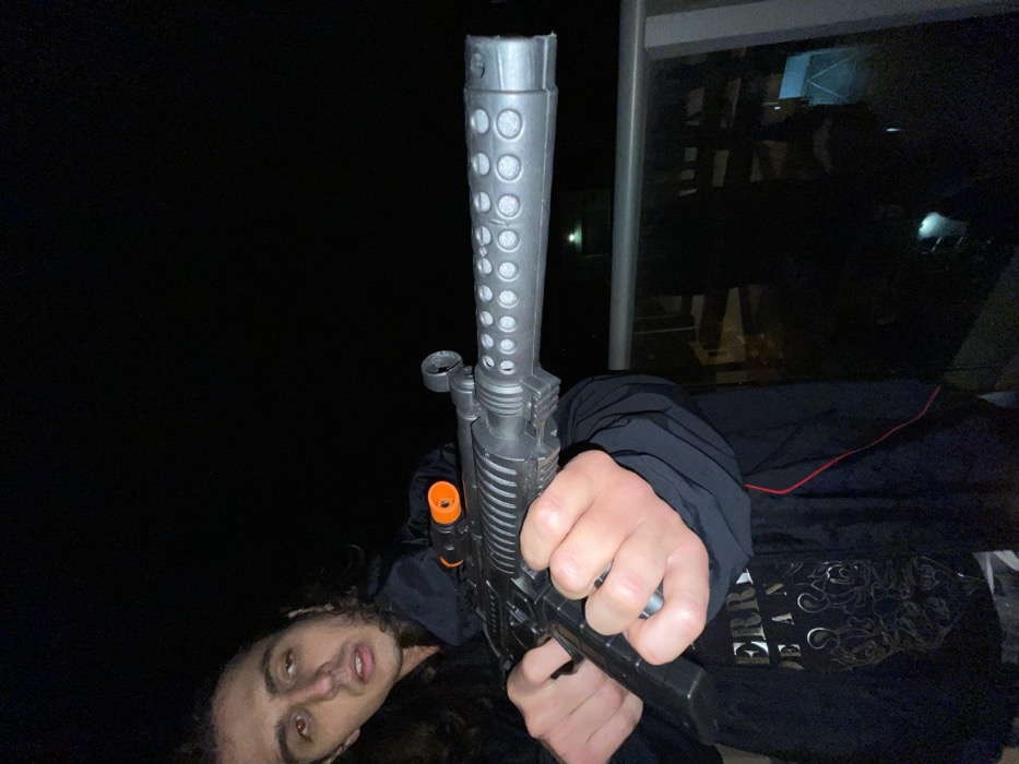 A male individual is depicted at night on a balcony in Tel-Aviv, Israel. The subject has long, dark, curly hair and wears a dark hooded jacket over a black t-shirt with a light-colored, ornate design. He is holding a grey and black object, resembling a firearm, with both hands in an extended, forward-pointing posture. The object features a perforated barrel shroud and an orange plastic sight element on its upper surface. A red-eye effect is present in the subject's left eye. The background consists of darkness with faint distant urban lights visible beyond a balcony railing constructed from metal and a transparent panel.