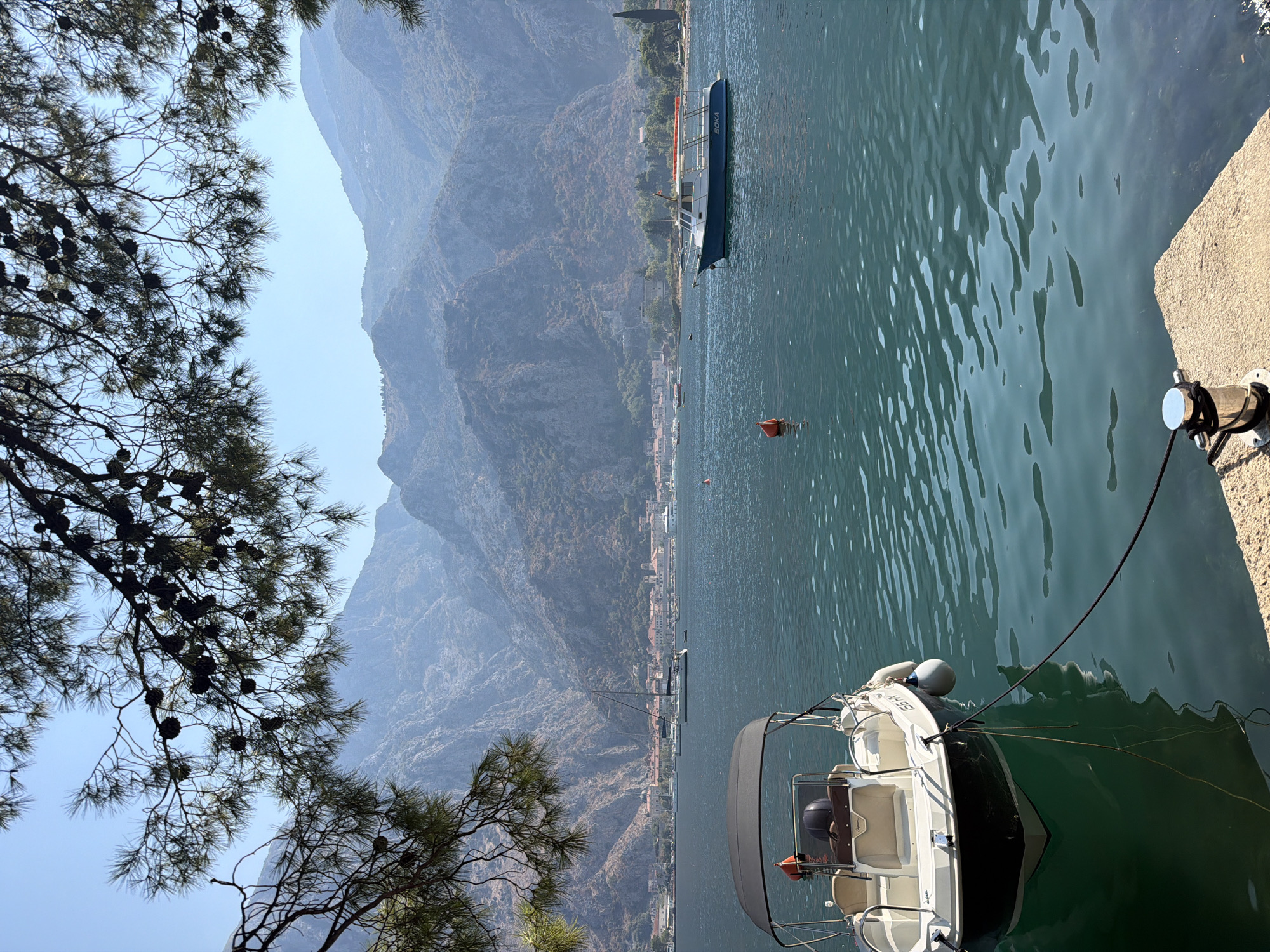 The image depicts a serene bay in Škaljari, Montenegro, with a clear blue sky and lush green mountains in the background. In the foreground, a white boat with a black canopy is moored near a concrete dock. The boat is tethered by a black rope to a silver bollard on the dock. Further out in the bay, another boat with a blue hull and a white superstructure, labeled "BOKA," is anchored. The water is calm and reflects the surrounding scenery. Pine branches with cones hang from the top of the frame, framing the picturesque view. The town of Škaljari is visible in the distance, nestled at the foot of the mountains. The overall scene conveys a sense of tranquility and natural beauty.