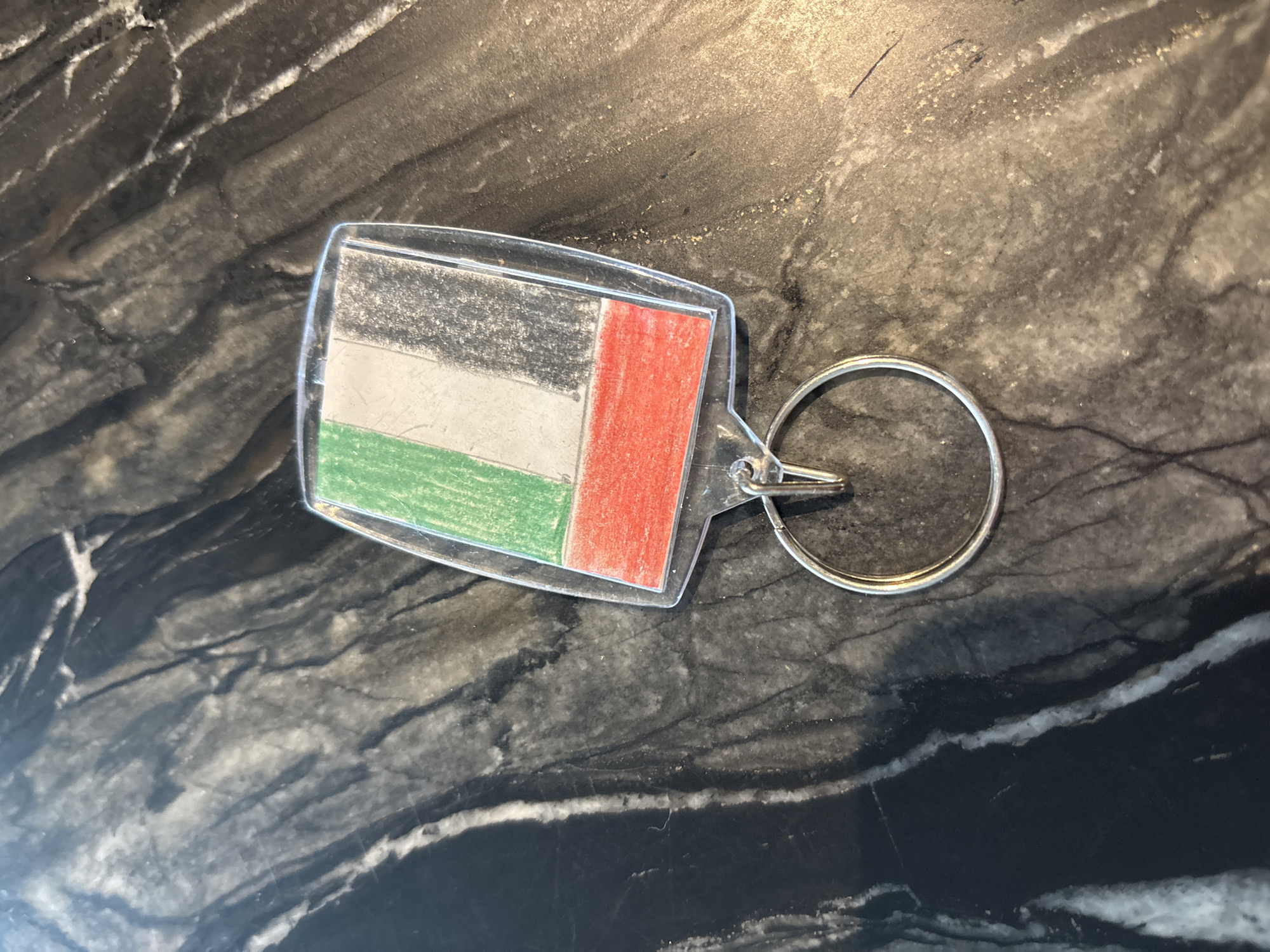 A close-up, top-down shot shows a United Arab Emirates flag keychain lying on a dark, marbled surface. The flag has vertical stripes of green on the left, white in the middle, and black on the right, with a horizontal red stripe at the bottom. The keychain is made of clear plastic and is attached to a silver keyring. The marbled surface is a mix of black, gray, and white, with a slightly reflective quality. No people are visible in the image. The scene appears to be indoors, possibly on a table or countertop. There are no discernible landmarks or environmental features that would indicate a specific location within Dubai.