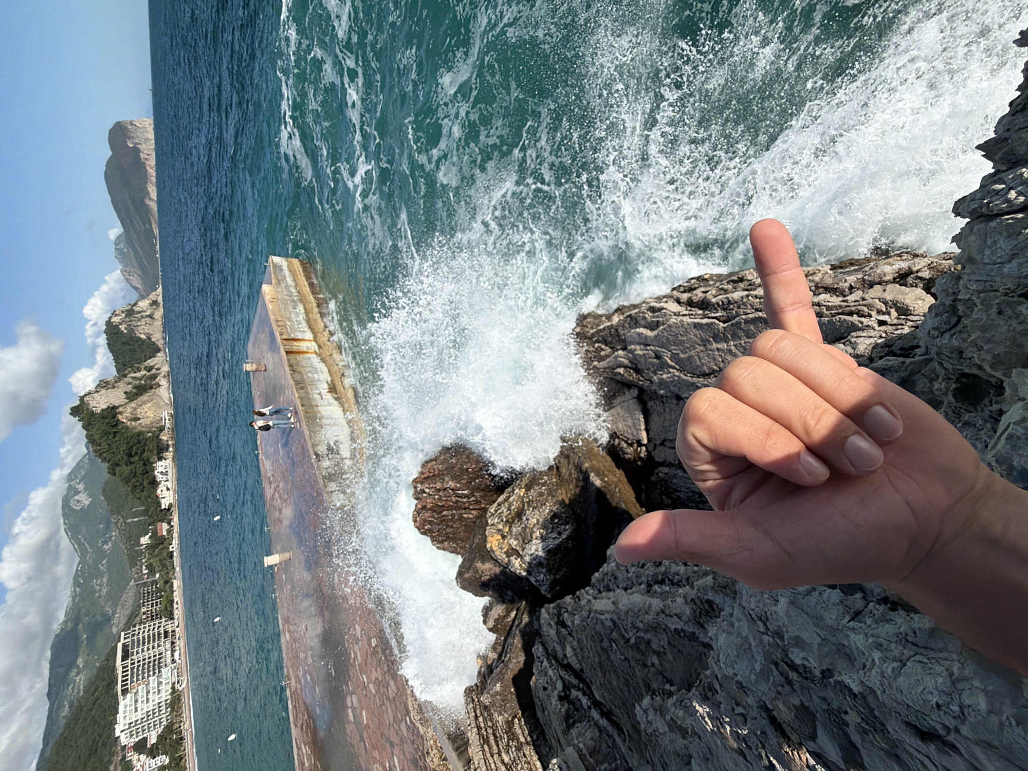 The image is a rotated, close-up view of the coastline in Petrovac centar, Montenegro. A human hand is in the foreground making a “hang loose” hand sign. The background shows the Adriatic sea and Petrovac's coastline. Waves are crashing against rocks. Two individuals are standing on a concrete pier to the left. Mountains with buildings extend across the image. The sky is partly cloudy.