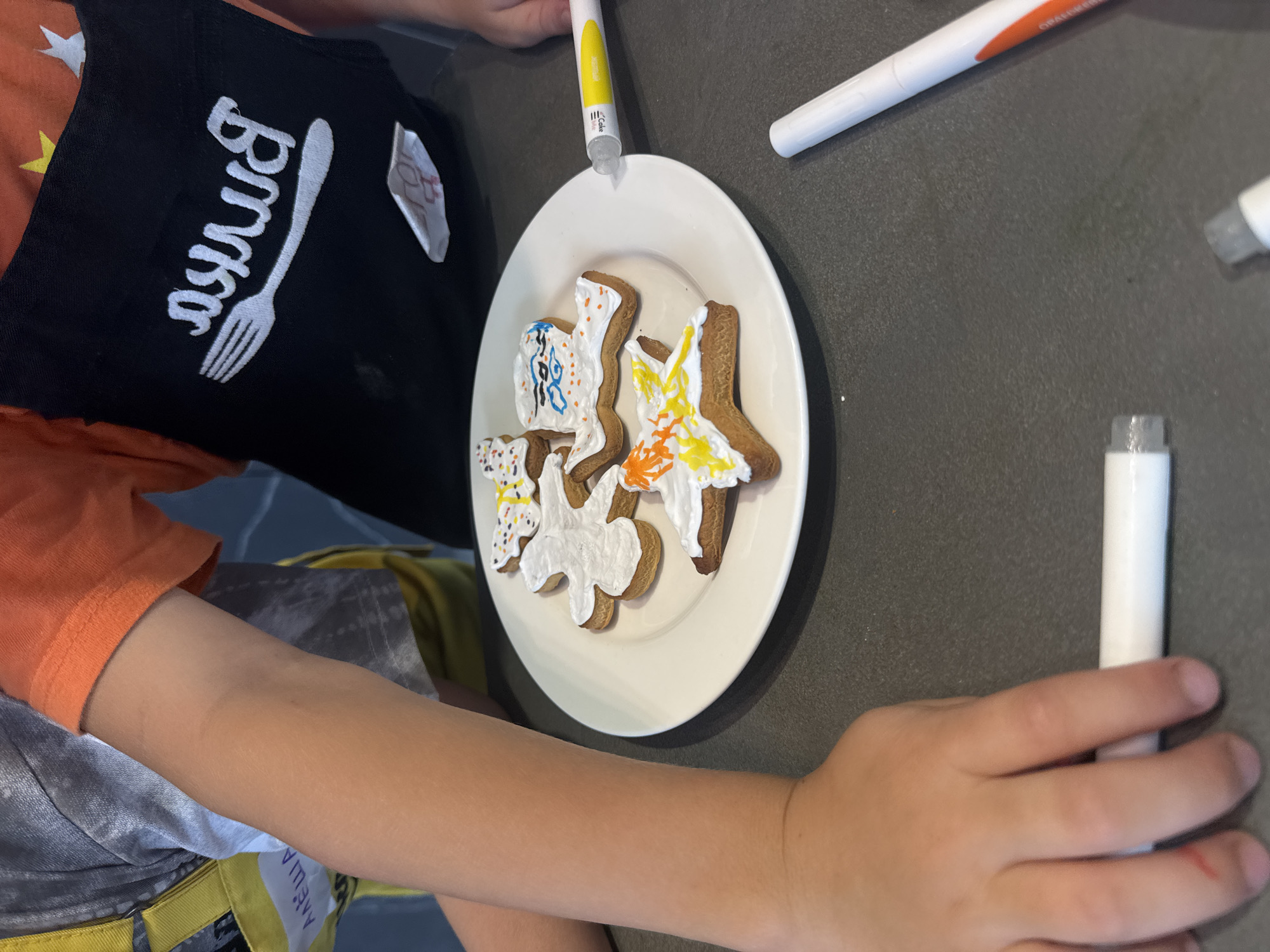 A child decorates gingerbread cookies on a white plate. The child wears an orange shirt and a black apron with white writing. The plate contains decorated gingerbread cookies shaped like a star, a person, and other shapes. There are several white and yellow pastry decorating pens surrounding the plate. The background is a gray surface. There is no indication of the location being Izhevsk, Russia.