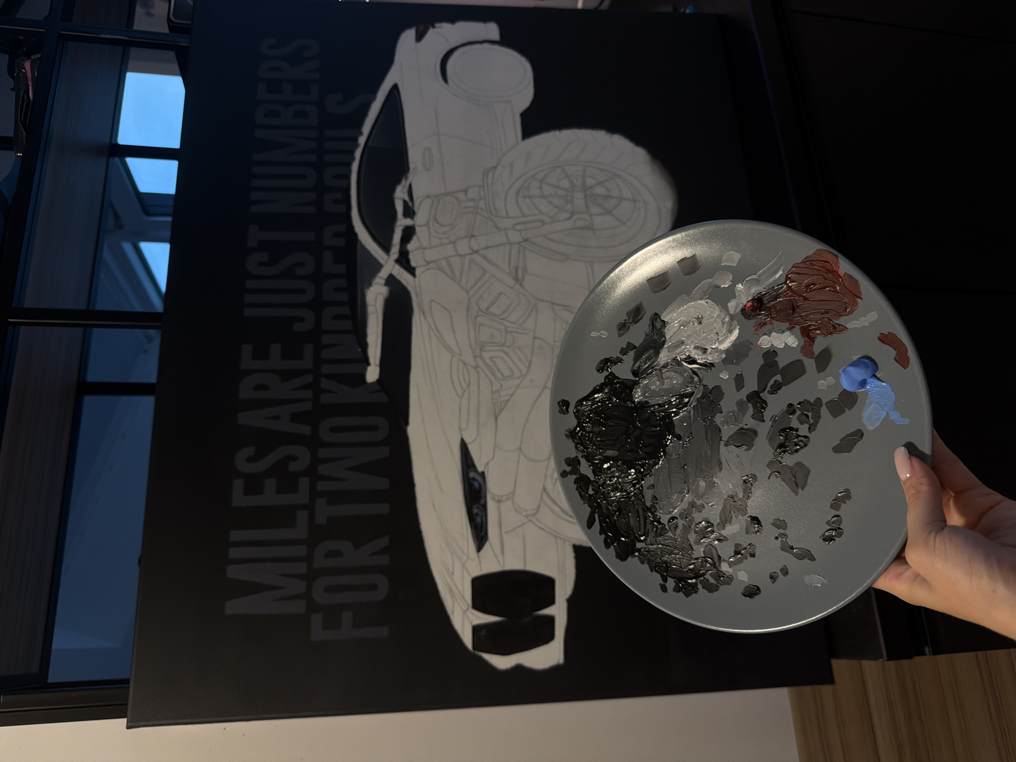 A person is holding a gray circular artist's palette covered in black, gray, white, brown, and blue paint, positioned over a black canvas leaning against an indeterminate structure in an indoor setting. The canvas has a white line drawing of a car and the gray text "MILES ARE JUST NUMBERS FOR TWO KINDRED SOULS" printed on it. A window with a black frame is visible in the background, revealing a bright, partly cloudy sky. The location is unknown.