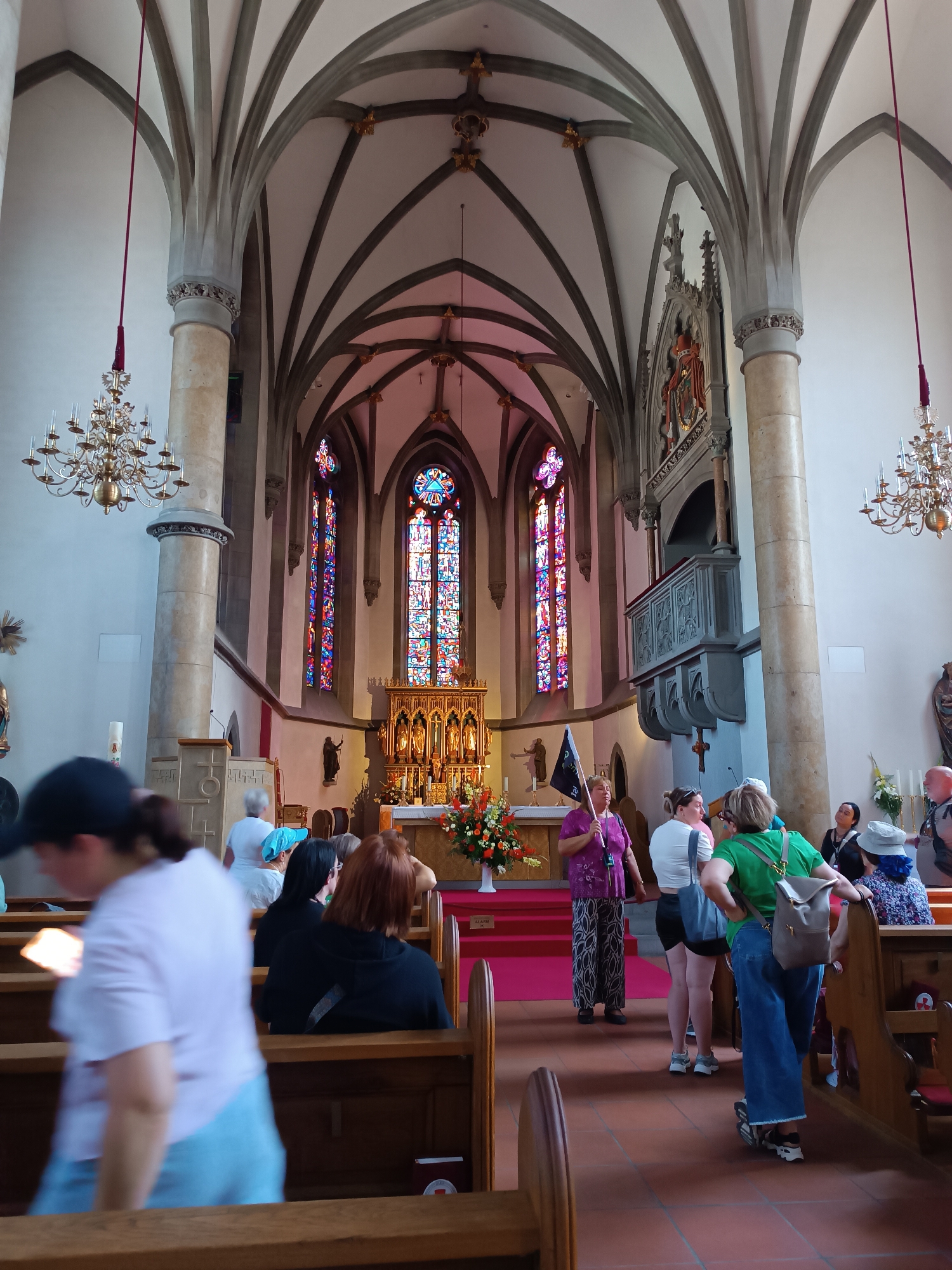 The image depicts the interior of a large, ornate church, likely a cathedral, with high vaulted ceilings and Gothic architectural elements. Stained glass windows with vibrant colors are prominent, illuminating the space. In the foreground, several people are visible, some seated in wooden pews and others standing and observing. A woman in a purple top and patterned pants holds a black flag, possibly as part of a tour or demonstration. The overall scene suggests a public gathering or visit within a significant religious building. There is no clear indication of the location being in Vaduz, Liechtenstein; it is a generic interior of a European church.
