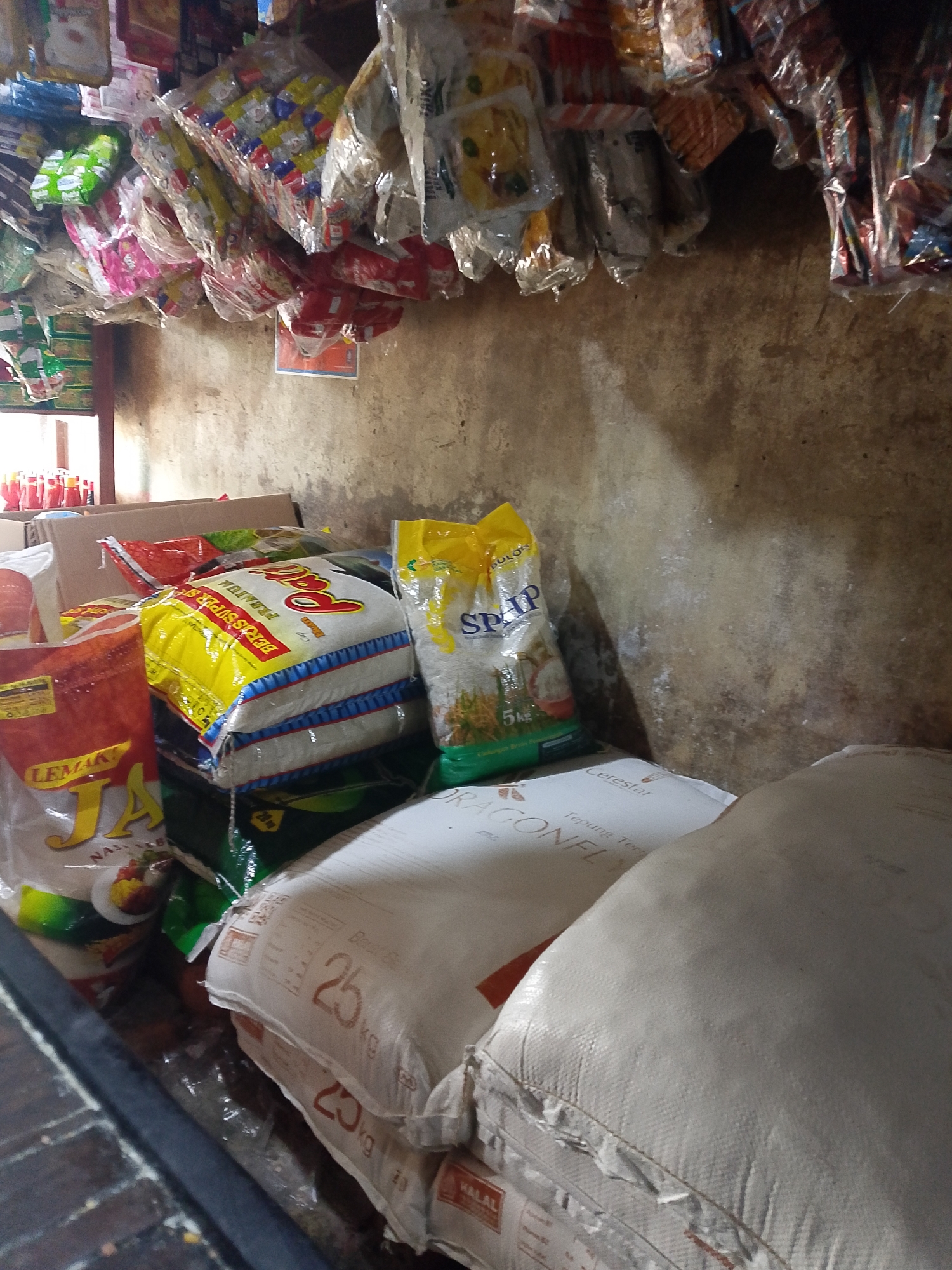 A dimly lit, rustic, interior scene depicts a cluttered shelf and counter space in what appears to be a small Indonesian convenience store or market. The wall behind the merchandise is rough and textured, with visible water stains and discoloration, suggesting it is an older building. The shelf above is laden with various bagged and packaged goods, many of which are colorful and appear to be food items or snacks.
Below the shelf, a collection of larger sacks and bags are stacked on the counter. Prominently displayed is a large yellow bag of rice labeled "Pati Beras Super SP". To its right, a yellow bag with "SDAP" branding is visible, also containing rice. In front of these, a red and orange bag with "LEMAKI JA" branding is partially visible. The foreground is dominated by two large, white woven sacks, one clearly marked with "25 KG" and the other with similar markings suggesting it is also a 25kg sack. The top sack has text that includes "Cerestar" and "DRAGONFLI". The overall impression is one of abundance and variety of staple goods. The natural light from a window on the left suggests the time of day is daytime. The scene is typical of numerous small, local businesses found throughout Palembang, Indonesia, where such goods are commonly sold.