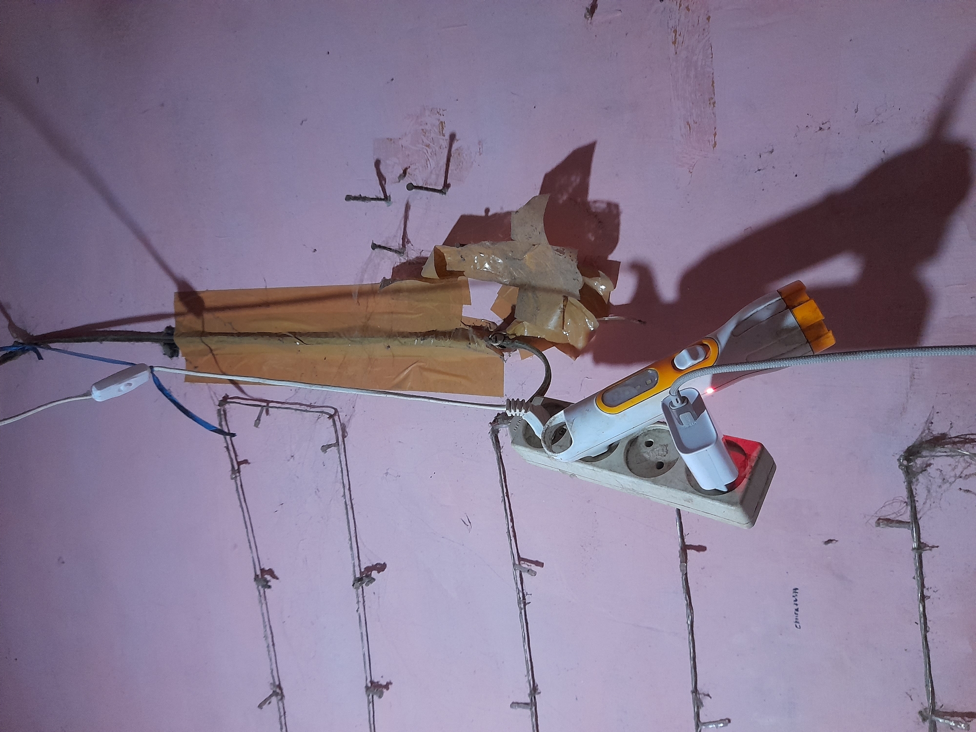 In Pulokulon, Indonesia, an overhead view captures a pink-toned wall with exposed wiring. A white power strip with three sockets is plugged into a wall outlet, and a white and orange device is plugged into the power strip. The device has a grey cord attached. The wall has several horizontal wires secured with small clips, and a piece of tan tape is affixed to the wall, holding a thicker cable. Nails protrude from the wall near the tape. The lighting casts a large shadow of the plugged-in device onto the wall.