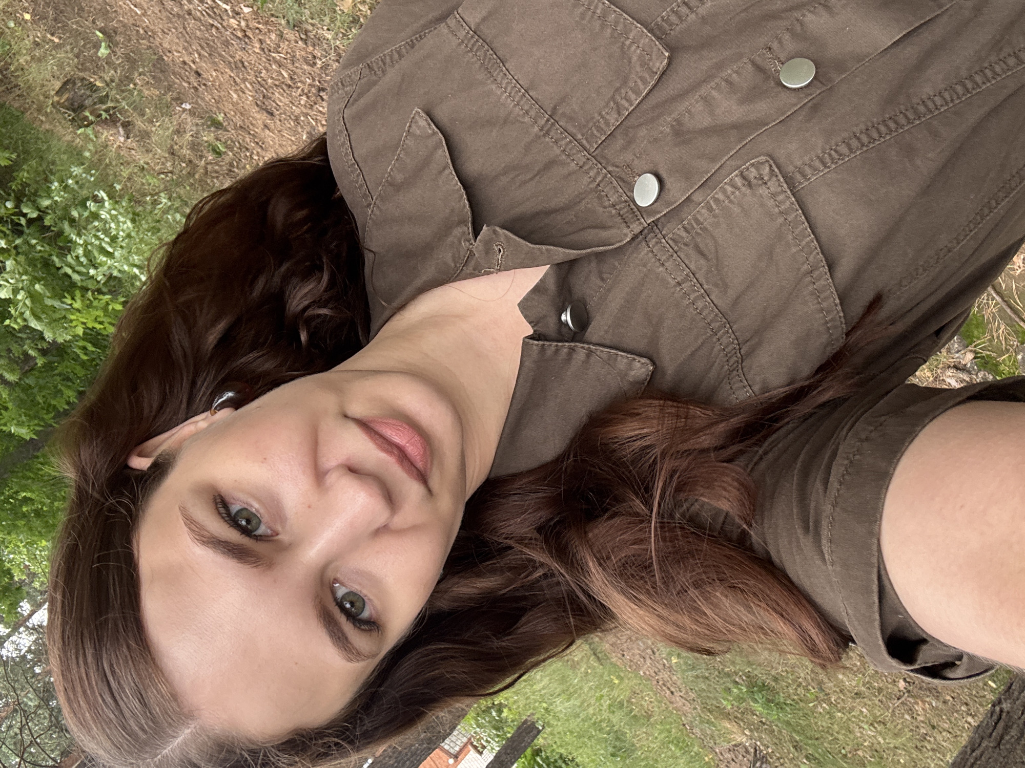 This media file is a close-up, portrait-style selfie of a young woman outdoors. She has long, wavy, dark brown hair and fair skin. Her eyes are green and her lips are lightly colored. She is wearing a dark olive-green jacket with metal buttons and stitched detailing. The background appears to be a natural setting with greenery and dry ground, suggesting a park or wooded area. The lighting is natural and diffuse, indicating it could be daytime, possibly overcast or late afternoon. There are no visible signs of text in the image. The woman's expression appears relaxed and content. The image is rotated 90 degrees clockwise.