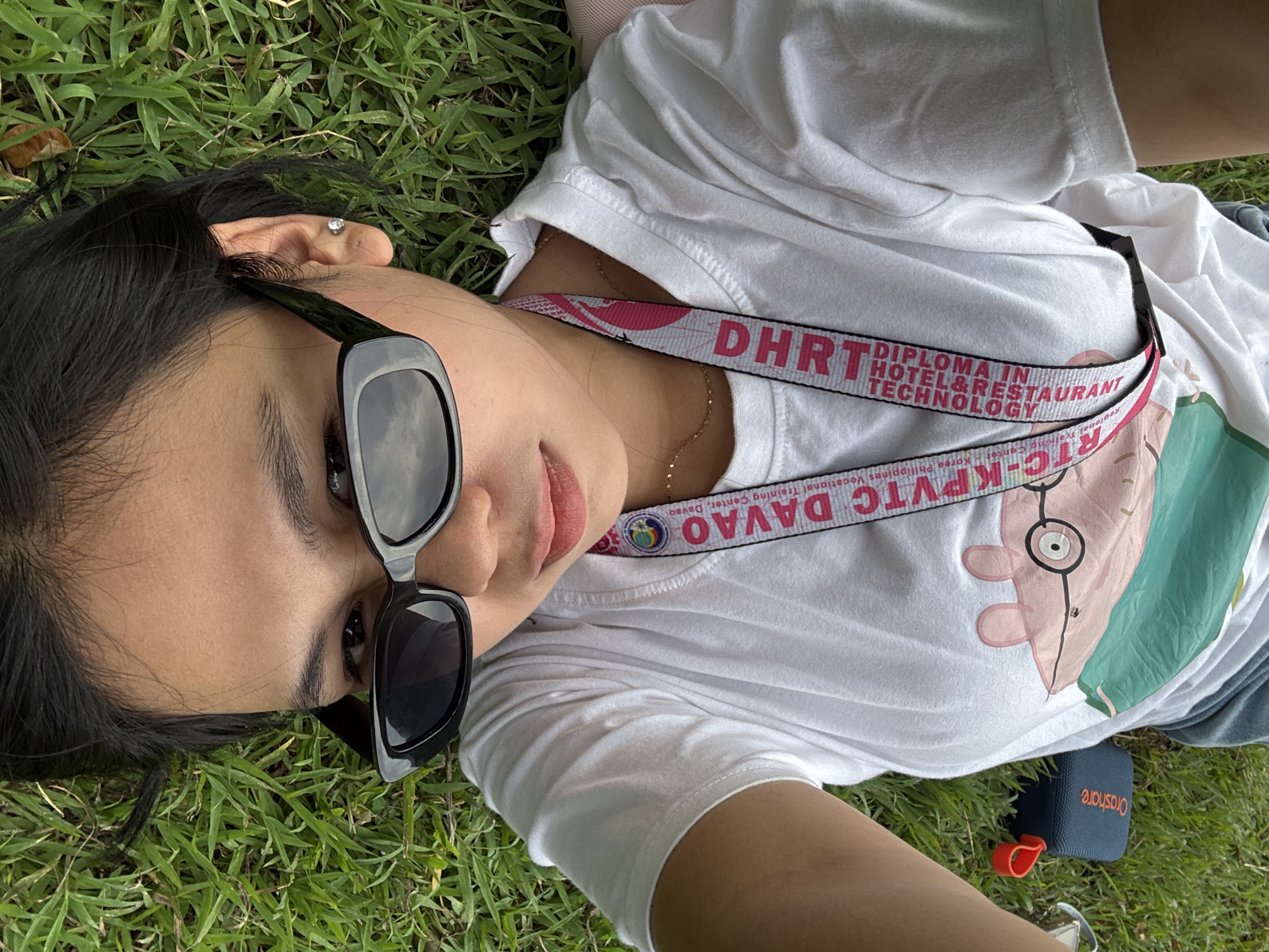 A young woman lies on a patch of green grass, looking up at the camera. She is wearing a white t-shirt with a graphic print on the front and a lanyard with text that reads "DHRT DIPLOMA IN HOTEL & RESTAURANT TECHNOLOGY" and "RTC-KPVTC DAVAO - Korea Philippines Vocational Training Center, Davao." She has on black-rimmed sunglasses and a small diamond earring. Her dark hair is spread out on the grass. A small, dark blue portable speaker with an orange strap sits on the grass near her leg. The lighting suggests it is daytime, possibly sunny, and the setting appears to be outdoors in a park or grassy area. The woman's expression is neutral, and the overall mood is relaxed.