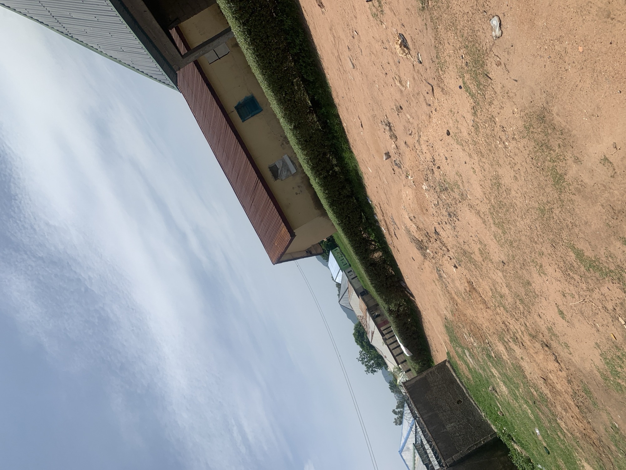 The image depicts an outdoor scene in Jauro Yinu, Nigeria. In the foreground, a dusty, reddish-brown dirt path occupies a significant portion of the lower half of the frame, with sparse patches of green grass growing through the soil. To the left, a section of a grey concrete wall with barbed wire is visible, adjacent to a grassy area.
In the mid-ground, a well-maintained green hedge runs horizontally across the image, separating the dirt path from a row of buildings. These buildings have light yellow or cream-colored walls and reddish-brown corrugated metal roofs. One building in the foreground has a blue-framed window and another with a partially covered window. Further in the distance, more buildings with similar roofing materials are visible, interspersed with trees and rolling hills.
The background is dominated by a pale blue sky with scattered white clouds, suggesting a partly cloudy day. The overall composition is angled, with the horizon line appearing tilted. There are no people or animals visible in the scene. The environment appears to be a semi-urban or rural residential area.