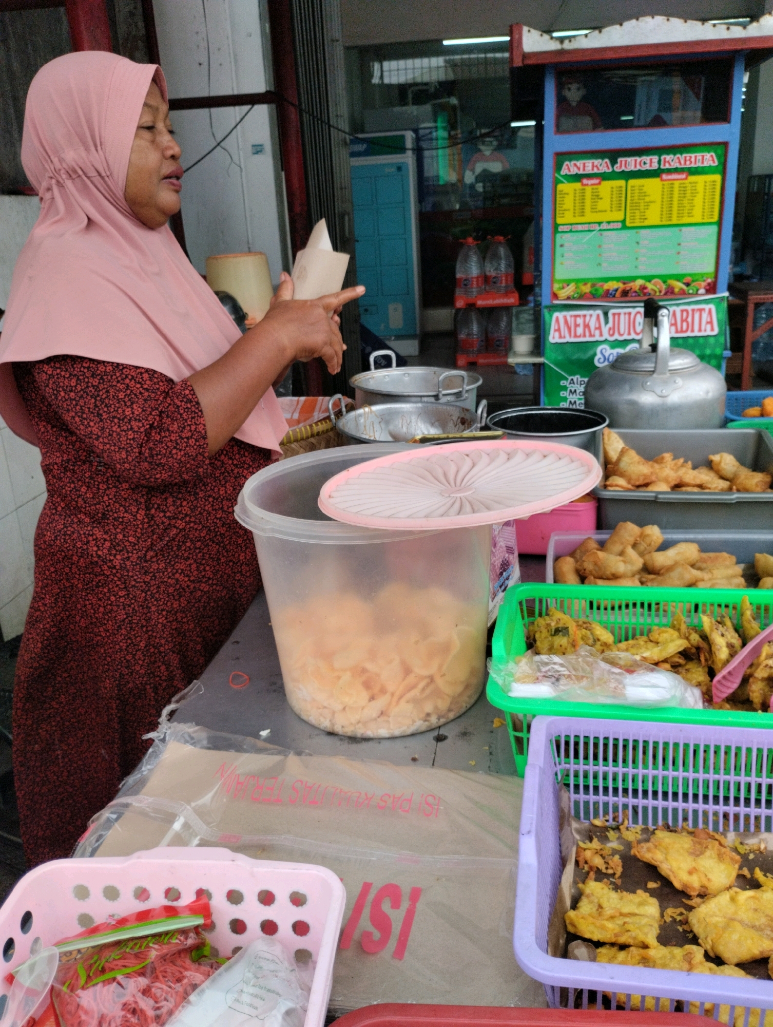 An outdoor stall selling juice and fried snacks is depicted in Bekasi, Indonesia. A woman wearing a pink hijab and a red floral dress is standing at the stall, holding a cone-shaped paper in her hands. She appears to be serving a customer, though no customer is visible in the frame. The stall features a menu board for "Aneka Juice Kabita," displaying various juice options and prices. Several bottles of drinks are stacked behind the counter. In front of the woman, there are containers of fried snacks, including what appear to be fried bananas or sweet potatoes, and other savory fried items. One large transparent plastic container on the counter holds light-colored, thinly sliced items, possibly chips or crackers. A pink plastic basket to the left contains red rubber bands and other items in plastic bags. A purple plastic basket and a green plastic basket, both filled with fried snacks, are also visible. The overall environment suggests a bustling street food setting.