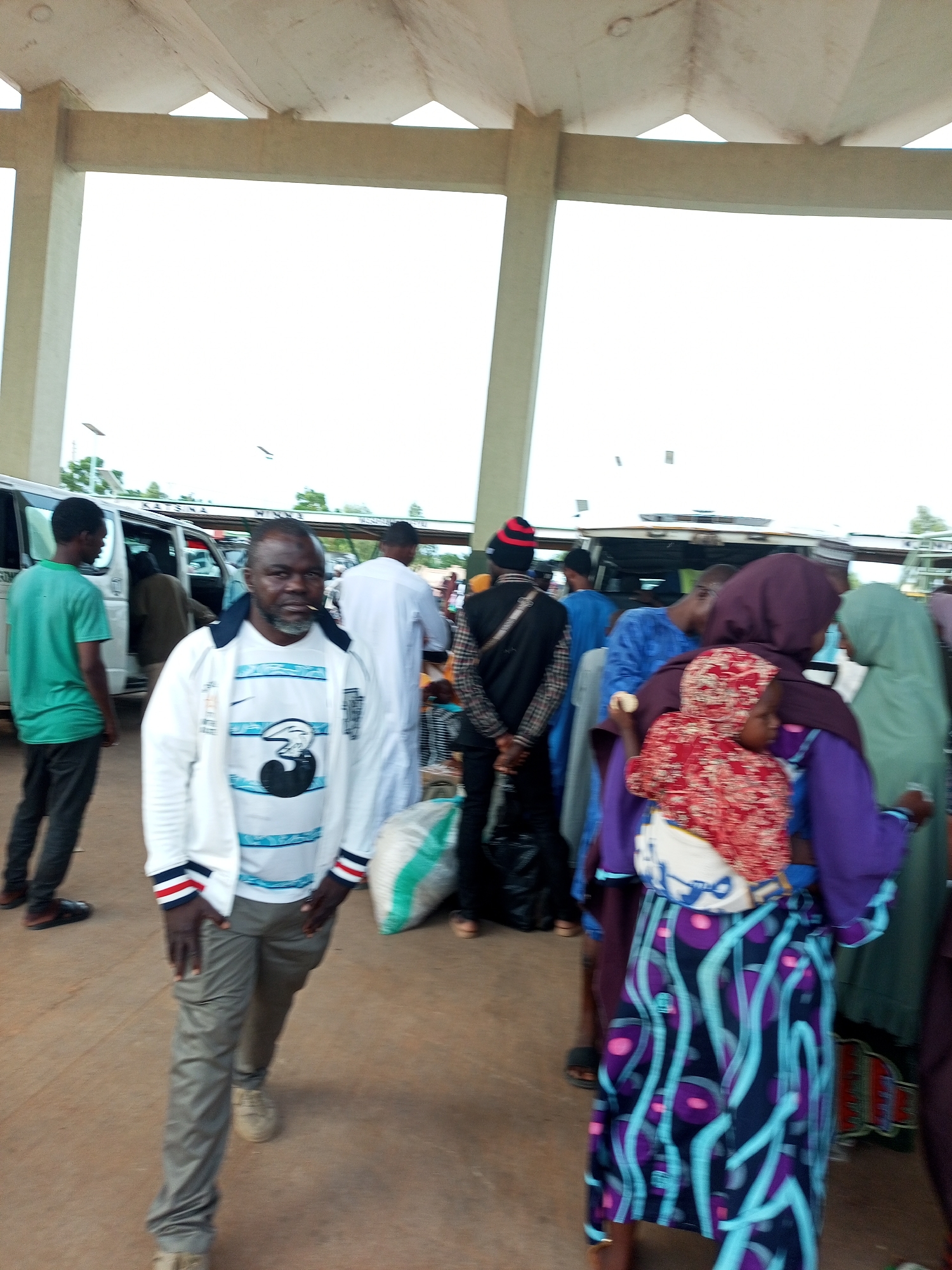 The image shows a group of people gathered under a covered structure with a concrete roof supported by pillars. The scene appears to be a transportation hub, possibly a bus park or a market area, in Gombe, Nigeria. In the foreground, a man in a white jacket with blue and red stripes on the sleeves and a white t-shirt with a graphic design is looking towards the camera. He is wearing grey pants and light-colored shoes. Behind him, several other individuals are visible, some standing near vans or buses. One person in a green shirt is walking away from the camera on the left. Another man in a white garment is standing further back on the right. A woman in a purple headscarf and a patterned blue and purple skirt is seen from behind, carrying a child. The ground is a reddish-brown dirt or pavement. The overall atmosphere suggests a busy public space.