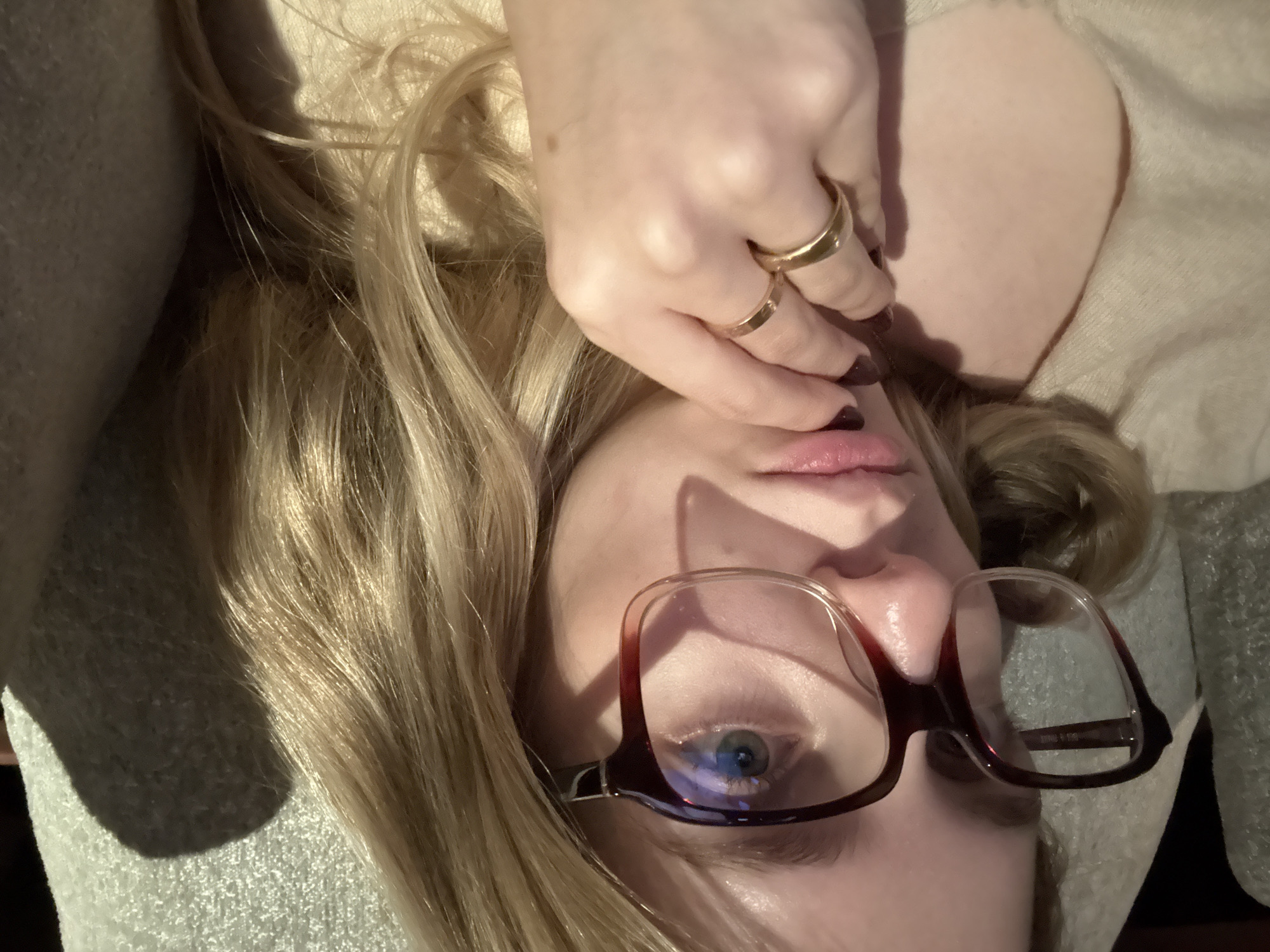 A close-up, angled portrait shows a woman lying down. Her head is angled towards the right, with her blonde hair spread around her head. She is wearing glasses with a dark red frame. Her right hand is covering part of her face. She is wearing two gold rings on her fingers and has dark nail polish on her fingernails. The background consists of beige clothing or fabric. The location is unknown.