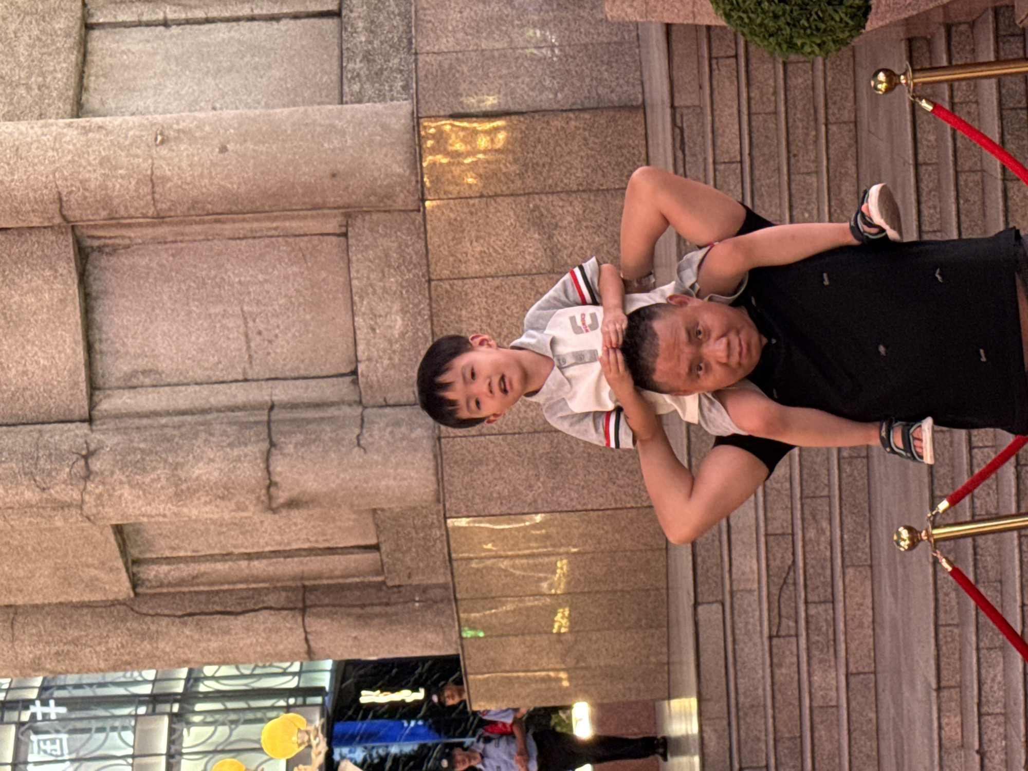 A father carrying his son on his shoulders stands on a set of outdoor stairs, with a red velvet rope barrier in the foreground. The father, a middle-aged man with short dark hair, wears a black polo shirt. His son, a young boy with black hair, sits on his shoulders, wearing a grey and white collared shirt with red and black trim and grey shorts. Behind them, a large stone building with textured panels forms the background. To the left of the father and son, two uniformed security guards stand near an entrance with a blue awning. The scene appears to be in the city of Yuzhong District, China.