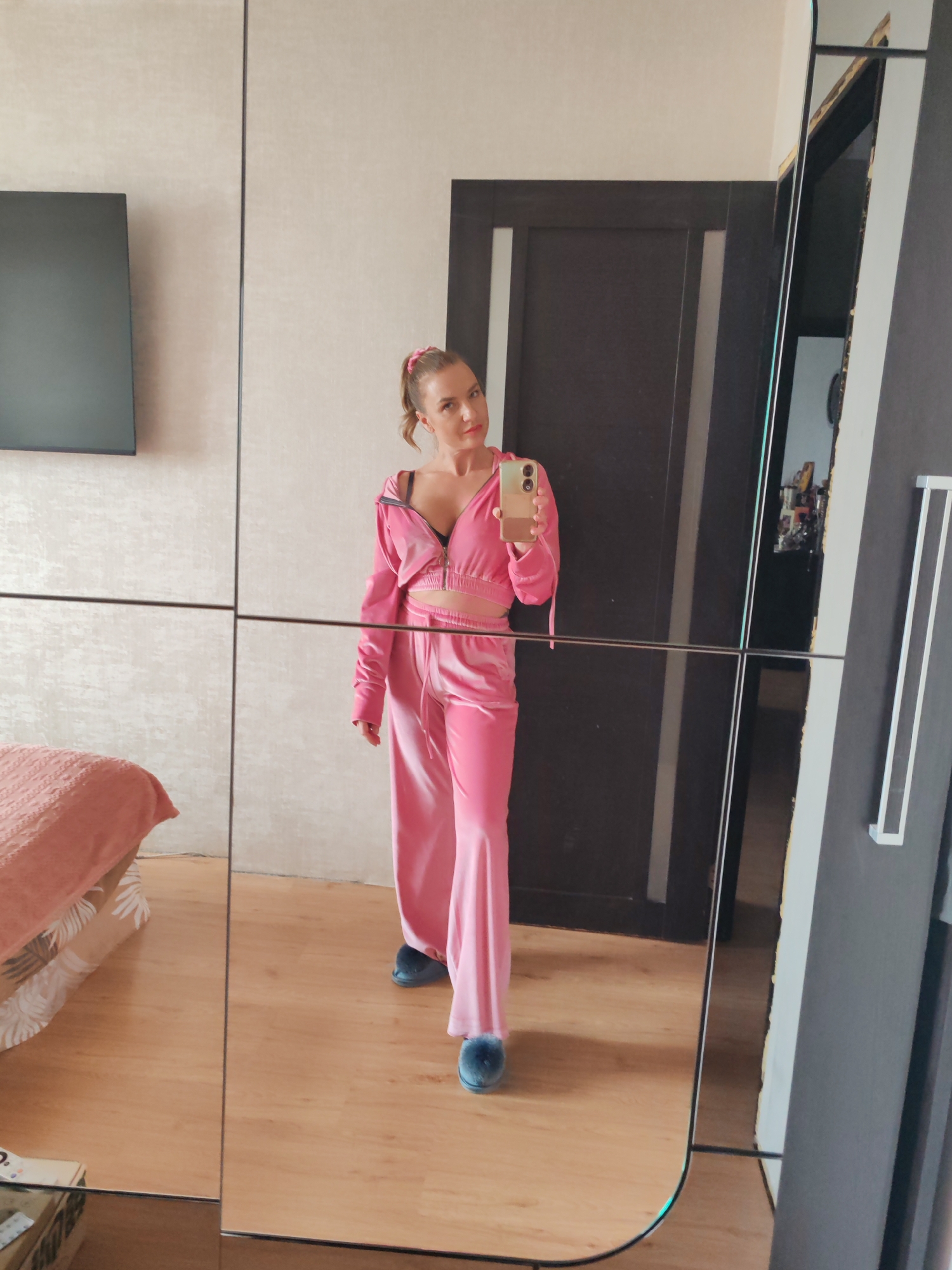 A woman stands in front of a full-length mirror, taking a selfie with her smartphone. She is wearing a pink velvet tracksuit and blue slippers. The background shows a bedroom interior with a bed, a TV screen mounted on the wall, and a door. The location is inferred to be a residential space in Petropavlovsk-Kamchatsky, Russia.