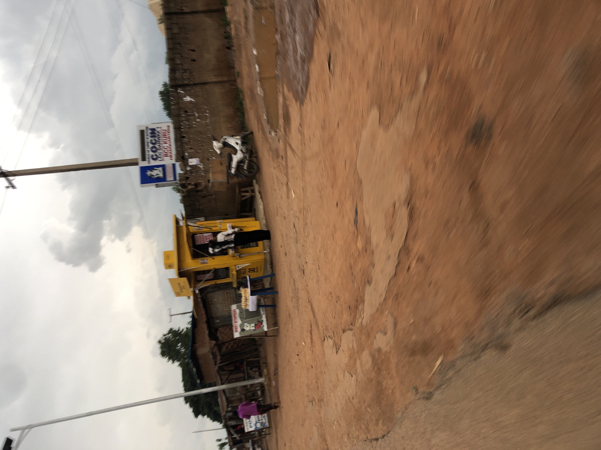 This is a medium-shot, eye-level, outdoor photograph taken in Vom, Nigeria. The scene depicts a street with a red dirt road and a partially paved area. On the right side of the road, there's a concrete wall with posters plastered on it. A white motorbike is parked in front of the wall. Adjacent to the wall is a yellow booth, possibly a small shop or service kiosk, with a person in black attire standing near it. In front of the booth, there's a blue table with items displayed on it. To the left of the booth, there are makeshift structures made of corrugated metal and wood. A person in a purple shirt is walking away from the camera towards these structures. In the background, a tall pole with electrical wires is visible, and trees are present. The sky is overcast with clouds.