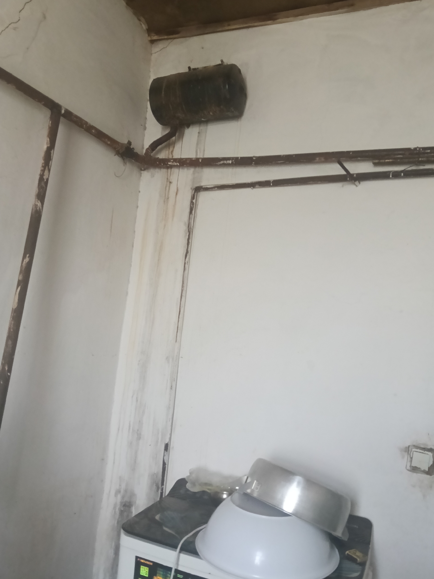 This is a forensic, matter-of-fact description of a scene in Altinkul, Uzbekistan. The image depicts an interior view of a room, likely a kitchen or utility area, with visible signs of age and disrepair.
Dominating the upper left of the frame is a dark, cylindrical object mounted high on the wall, possibly a water heater or storage tank. It is connected to a network of rusty, brown pipes that run horizontally and vertically along the walls and ceiling. These pipes show significant signs of corrosion and rust, suggesting long-term exposure to moisture or neglect.
The walls are painted a light color, but they are marred by extensive water stains and streaks, particularly running down from the pipework and the ceiling. Some areas of the wall appear cracked. The ceiling itself is also damaged, with visible cracks and discolored sections. A wooden beam is partially visible along the top edge of the image, also showing signs of wear.
In the lower part of the image, a washing machine is partially visible, with a label that reads "PREMIER." On top of the washing machine are two kitchen bowls, one large white plastic bowl and a smaller, shiny metallic bowl placed on top of it.
The overall impression is one of an aged, possibly disused or poorly maintained space. There are no people or animals visible in the image, and no active events are occurring. The scene is static and presents a condition assessment of the environment.