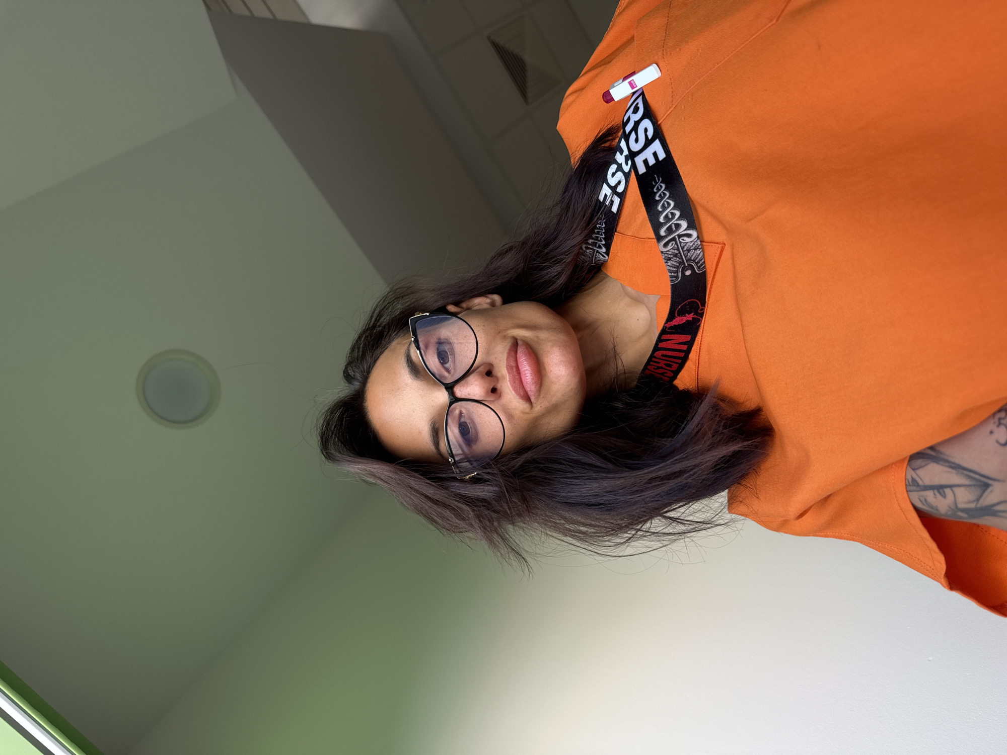 A woman wearing an orange scrub top and glasses is looking up at the camera. She has dark hair and is wearing a lanyard that says "NURSE". She has a tattoo on her arm. The background appears to be an indoor medical setting, possibly a hospital or clinic. The image does not provide any specific location information in Pleven, Bulgaria.