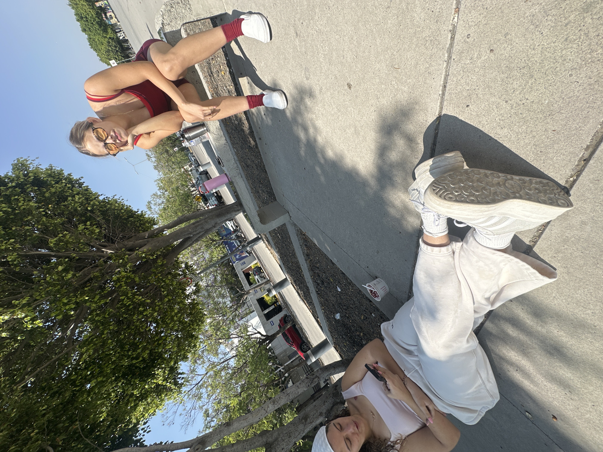 Two women are present in the image. One is in a red crop top and shorts with white sneakers and red socks, wearing sunglasses. She is sitting on a sidewalk with her legs crossed, turned towards the camera. The other is lying down on the sidewalk, wearing a white tank top and sweatpants with white sneakers. She is looking at her cell phone. They are both outside in what appears to be a sunny urban environment. In the background, there are cars parked along the side of the road, some trees and other buildings. Based on the direction of the trees and buildings, it seems that the photo may have been taken near a street or park. It is not possible to confirm the location in Puebla, Mexico based on the visual information available in the image.