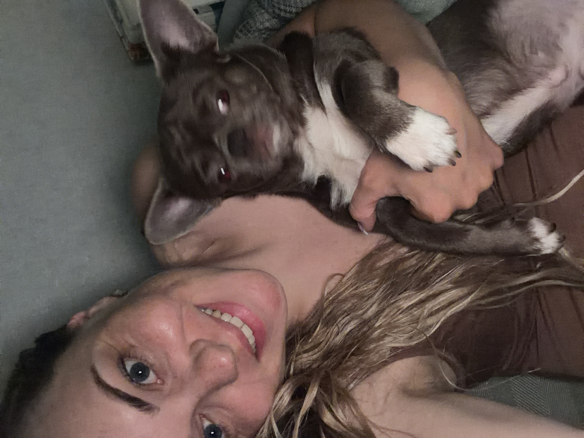 A woman with fair skin, light blue eyes, and blonde hair is lying down and smiling at the camera. She is holding a brown and white dog in her arms. The dog appears to be moving, as its face is slightly blurred. The background appears to be a couch or bed. There is no information in the image to identify a specific location or to verify that it is in the city of Votkinsk, Russia.