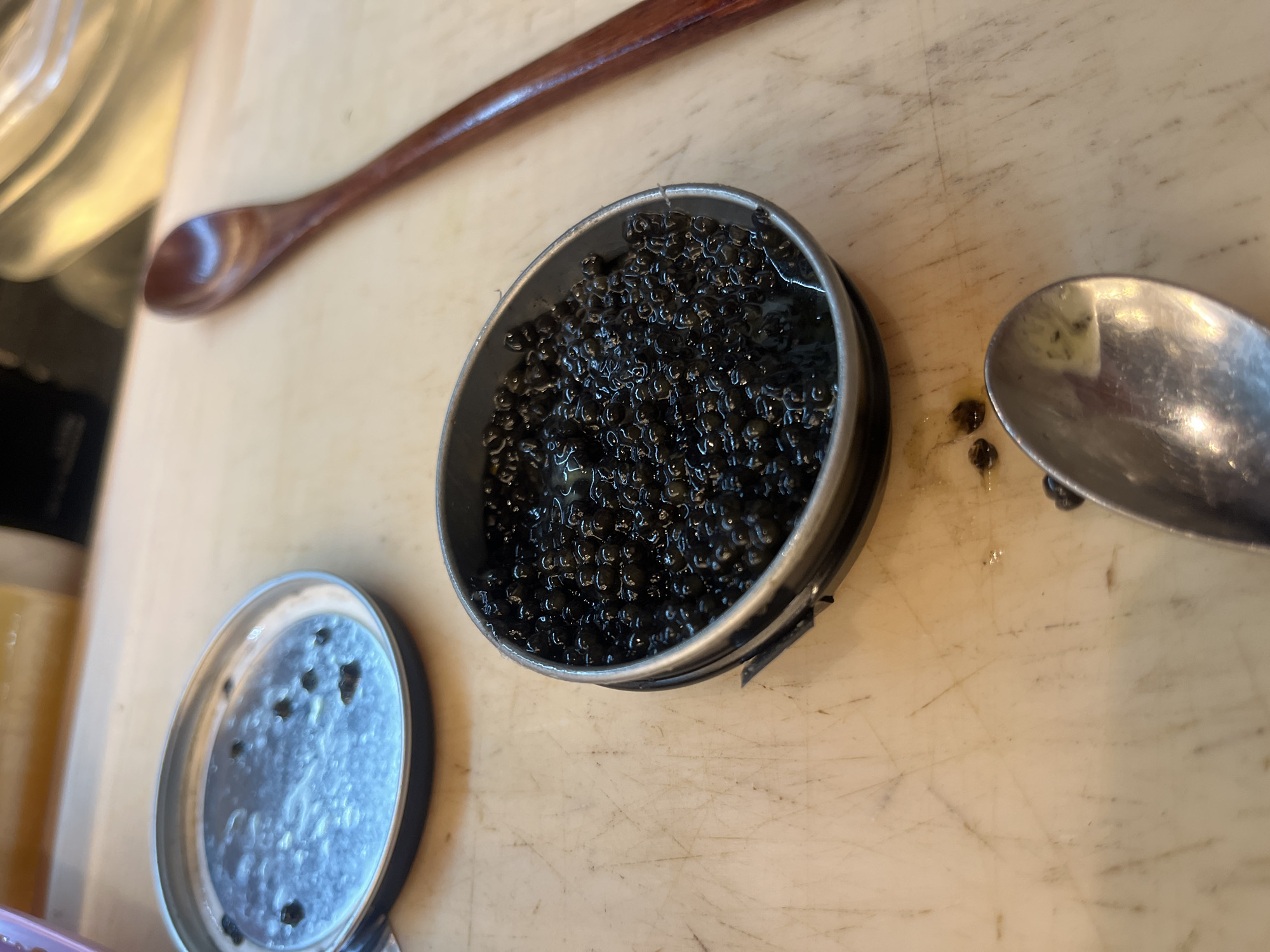 An overhead, slightly angled shot captures an open, round metallic tin brimming with glistening, dark black caviar. The tin is situated on a light-colored surface, possibly a well-used wooden or plastic cutting board or countertop, which exhibits numerous scratches and marks. To the left of the caviar tin, its circular lid lies detached, showing a few small, dark specks. A slender, polished dark brown wooden spoon rests diagonally in the upper left portion of the frame, partially out of focus. On the right, a silver metal spoon, likely a teaspoon, is positioned next to the caviar tin. A few individual caviar eggs are visible on the spoon's surface and scattered on the countertop nearby, suggesting recent handling or serving. The scene depicts a moment of food preparation or serving, likely in a kitchen or dining area in Sofia, Bulgaria. The lighting is even, suggesting an indoor environment. There is no visible text.