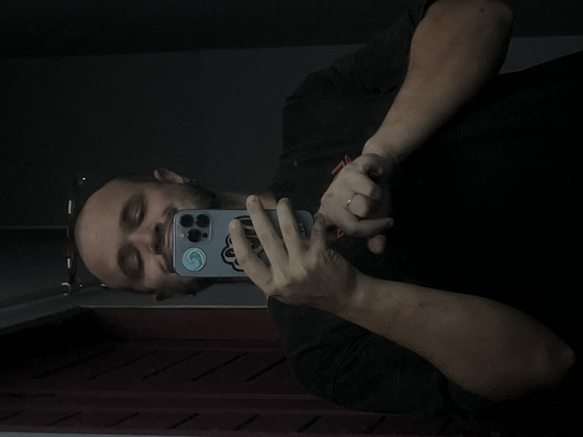 A man with a shaved head and stubble is taking a mirror selfie. He is wearing a black t-shirt. He is holding a light blue smartphone in his right hand, with his left hand resting on his hip. A ring is visible on his left ring finger. The background is dark, with a vertical red object on the left side of the image. The overall lighting is dim.