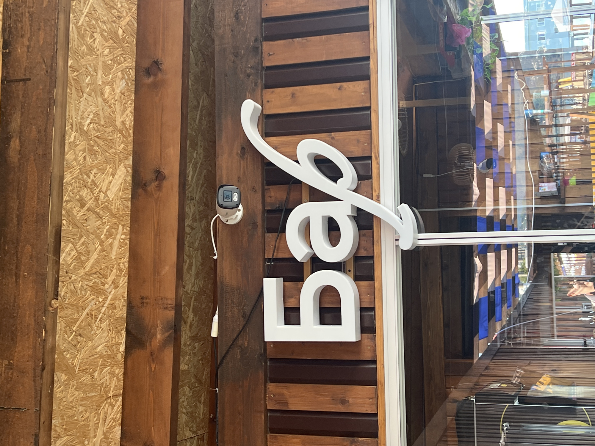 In Sochi, Russia, a white three-dimensional sign reading "Бар" (Bar) is mounted on a wooden wall. The letters are large and stylized, with the first letter "Б" being block-like and the other letters forming a cursive script. A security camera is visible mounted above and to the left of the sign. The wall behind the sign is constructed of vertical wooden planks, alternating between a lighter and darker shade of brown. Above this wall, there are horizontal wooden beams and a section of particle board. Below the sign, a glass storefront reveals the interior of the establishment, which appears to be a bar or cafe, with tables, chairs, and decor visible. The overall scene depicts the exterior signage of a business in Sochi.