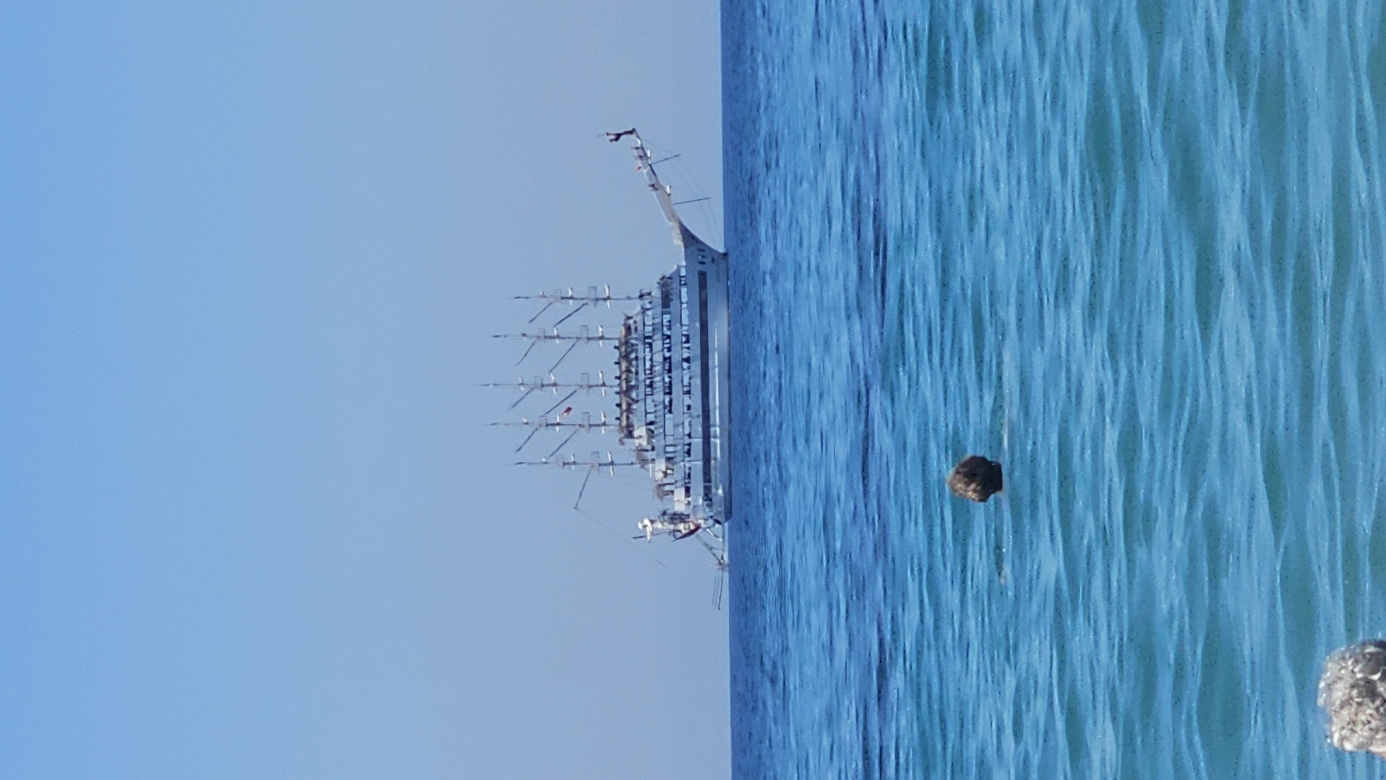 An image shows a large, white, multi-masted sailing ship on the blue sea under a clear blue sky. The ship has four masts and appears to be a cruise ship or a historical replica. In the foreground, a person's head and shoulders are visible just above the surface of the water, swimming. The water is a vibrant blue with ripples. The image is taken from the perspective of someone in the water. The location is the sea off the coast of Alanya, Turkey.