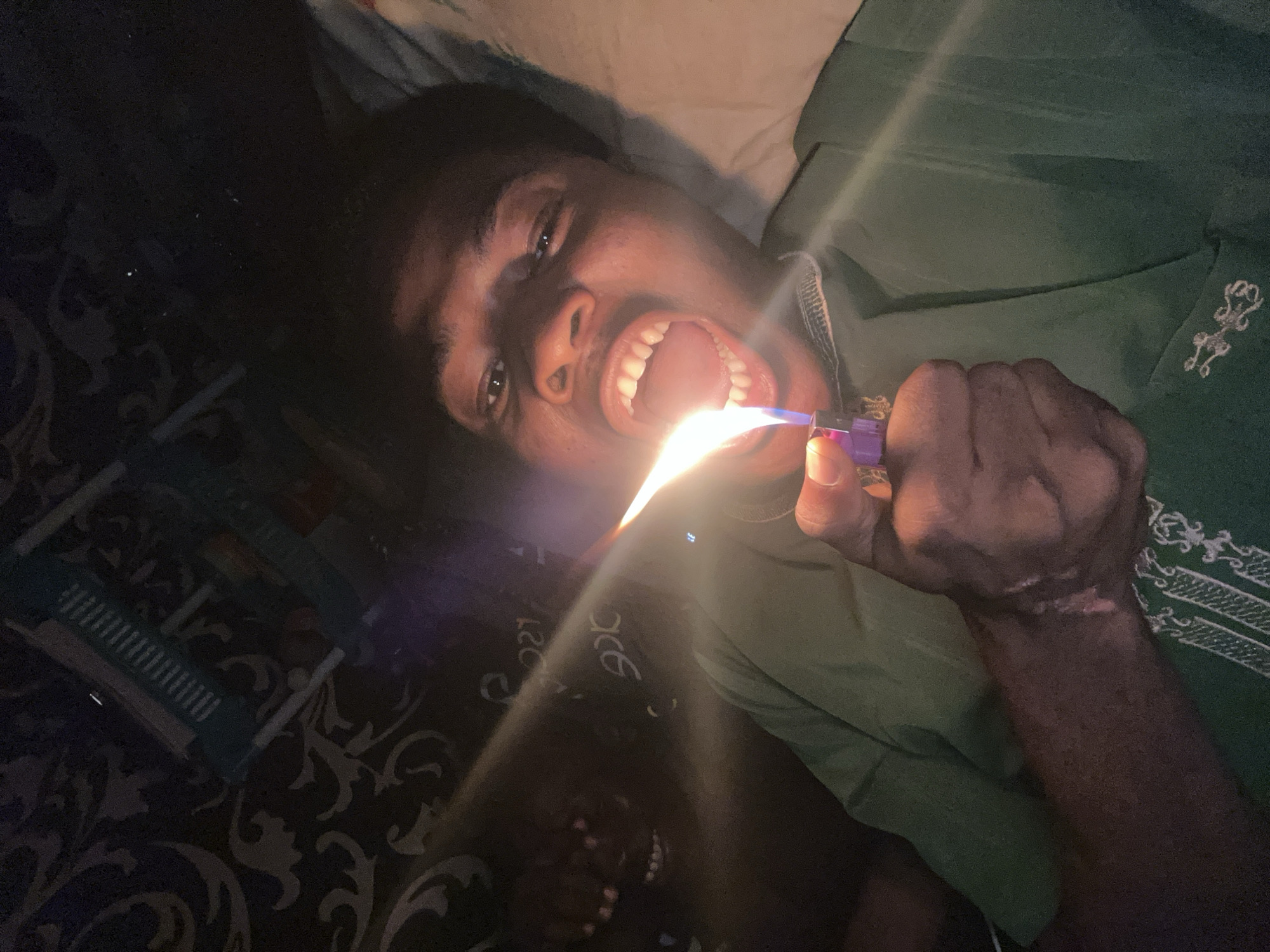 A dark-skinned young person with their mouth wide open is lying down and holding a purple lighter to their teeth. A bright flame erupts from the lighter, illuminating their teeth and the inside of their mouth. The person is wearing a green shirt with an embroidered design on the chest. They have dark hair and are looking directly at the camera with a somewhat surprised or excited expression. The background is dark and appears to be a patterned fabric, possibly a bedspread. The lighting is dramatic, with the flame casting strong shadows. Some text is visible on the patterned fabric in white, appearing to be stylized lettering. The context provided indicates the location is Ihiagwa, Nigeria. The image has a dark, indoor feel, suggesting it was taken at night or in a dimly lit room.