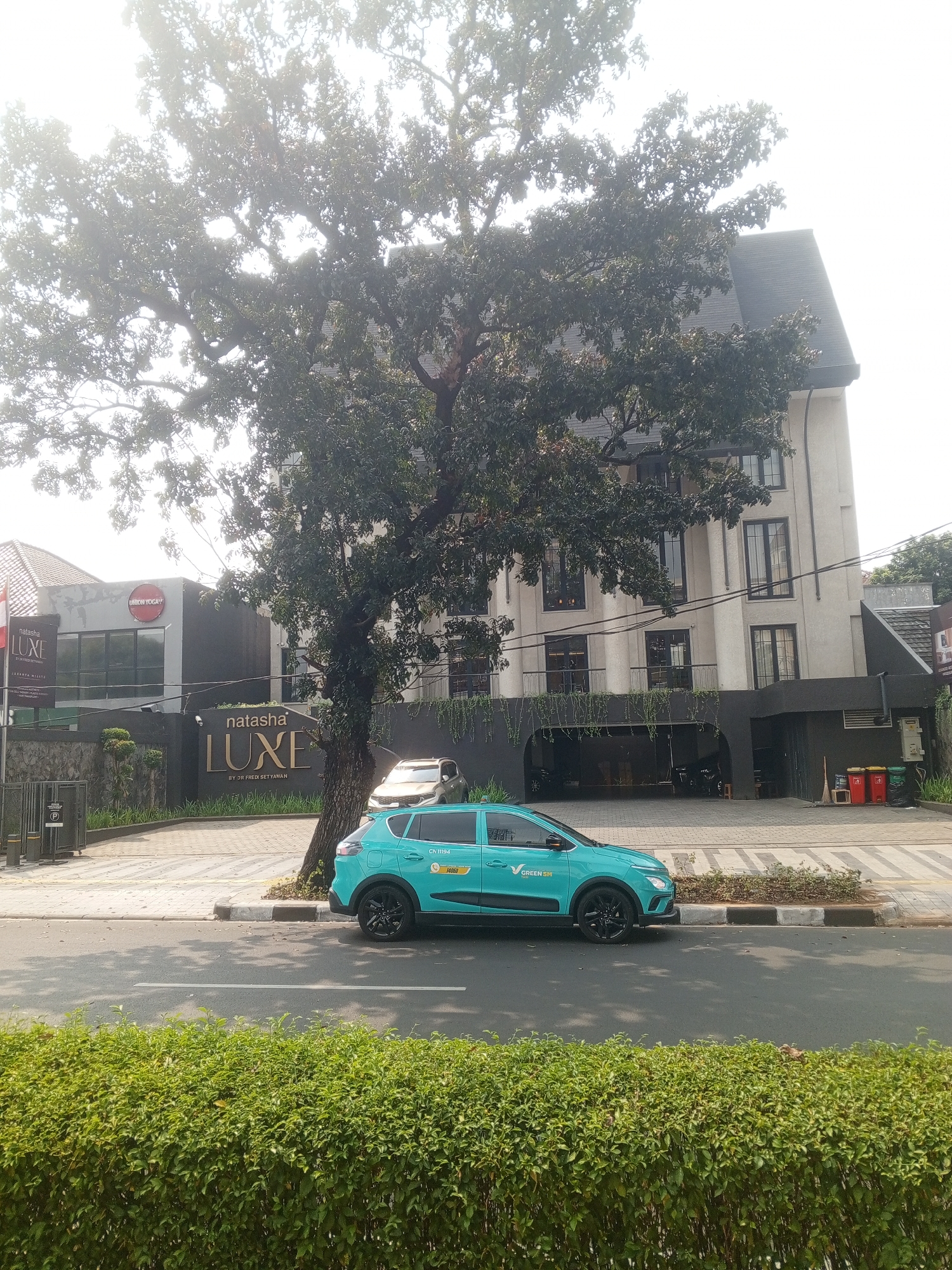 The image shows a street view in South Jakarta, Indonesia, featuring a modern building with a prominent "natasha LUXE" sign. A bright teal electric vehicle is parked on the side of the road, with the "GREEN SM" logo visible on its door, suggesting it is part of a ride-sharing or delivery service. A large tree with dense foliage stands between the road and the building, partially obscuring the view. The building appears to be a commercial establishment, possibly a beauty salon or clinic, given the "LUXE" branding. The street is paved, with a sidewalk and a neatly trimmed green hedge lining the foreground. Several trash bins are visible near the entrance of the building. The sky is bright, indicating daytime.