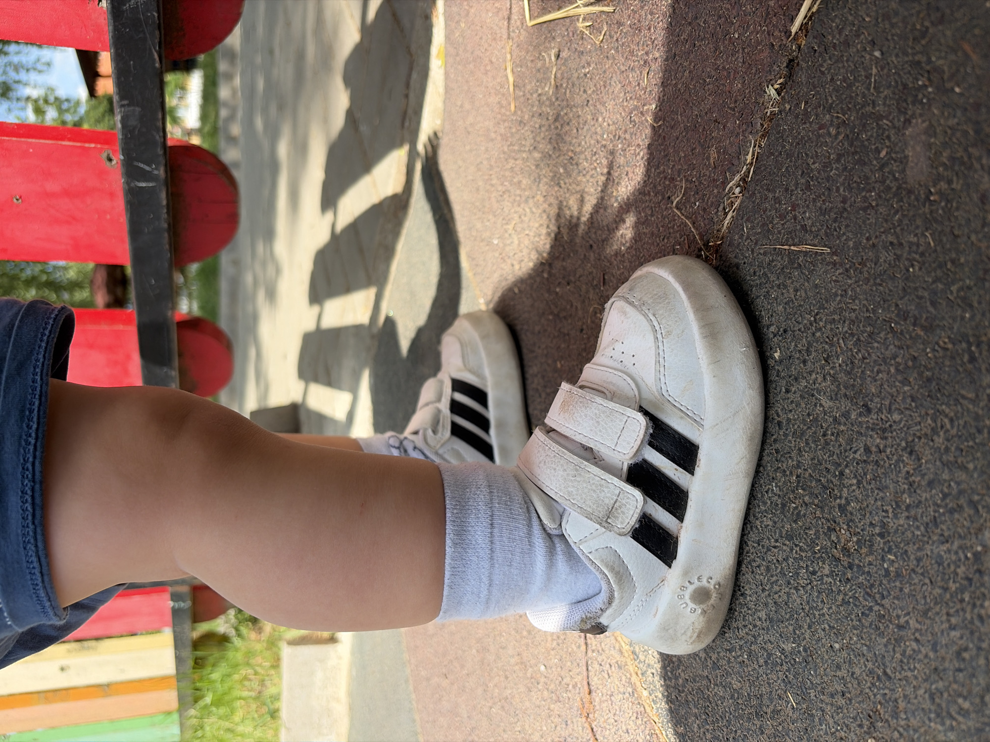 A photograph taken outdoors in Svetovrachene, Bulgaria, presents a partial view of a child. Only the child's left leg is visible from the upper thigh down, clad in a dark blue garment, a white sock, and a white sneaker with black stripes. The child is positioned next to a structure comprised of red and black elements on the left side of the frame. The background reveals a concrete or paved surface partially illuminated by sunlight casting shadows, adjacent to a dark rough surface scattered with light-colored debris.