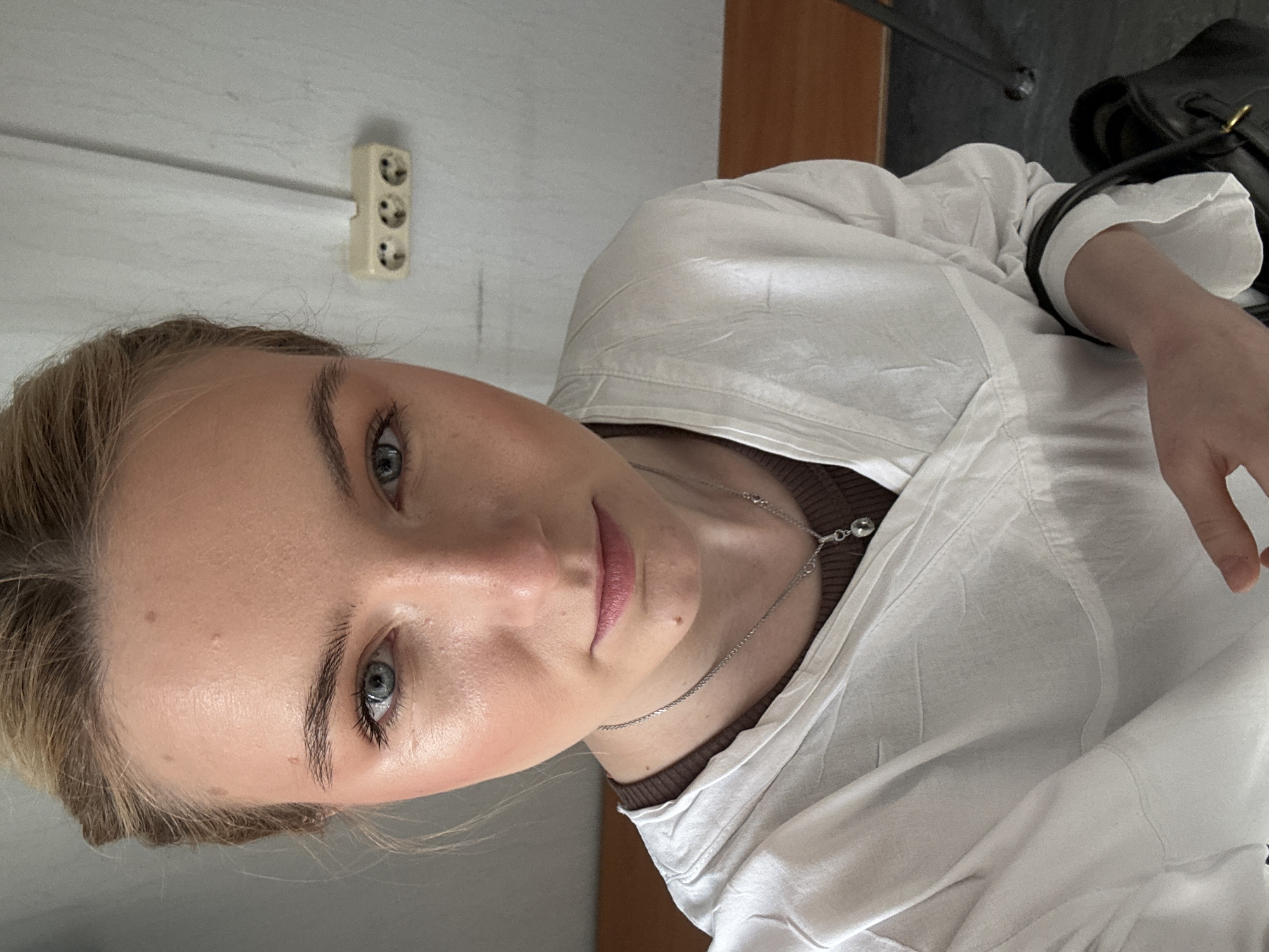 A close-up, slightly rotated image shows a fair-skinned woman with light brown hair and blue eyes. She wears a white button-down shirt over a brown top. A silver necklace adorns her neck. The background includes a light gray wall with a European-style electrical outlet, along with a glimpse of a wooden desk and a black bag. This image does not provide enough information to accurately confirm that it was taken in Saint Petersburg, Russia. More context would be needed.