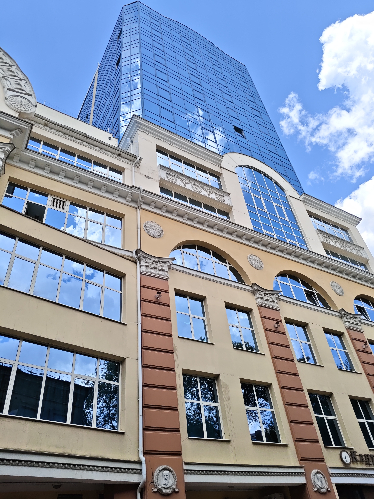 The image shows a low-angle view of a modern glass skyscraper reflecting the sky and clouds, towering over a traditional building with decorative architectural elements. The traditional building has a light beige facade with ornate white detailing around the windows and cornices. It also features a section with reddish-brown decorative pillars and framed panels. Several windows with white frames are visible on the traditional building, reflecting the blue sky and white clouds. The skyscraper, located in Rostov-on-Don, Russia, has a dark blue glass facade that creates a striking contrast with the older architecture. The sky is bright blue with scattered white clouds, indicating daytime. The image captures the juxtaposition of old and new architectural styles in an urban environment.