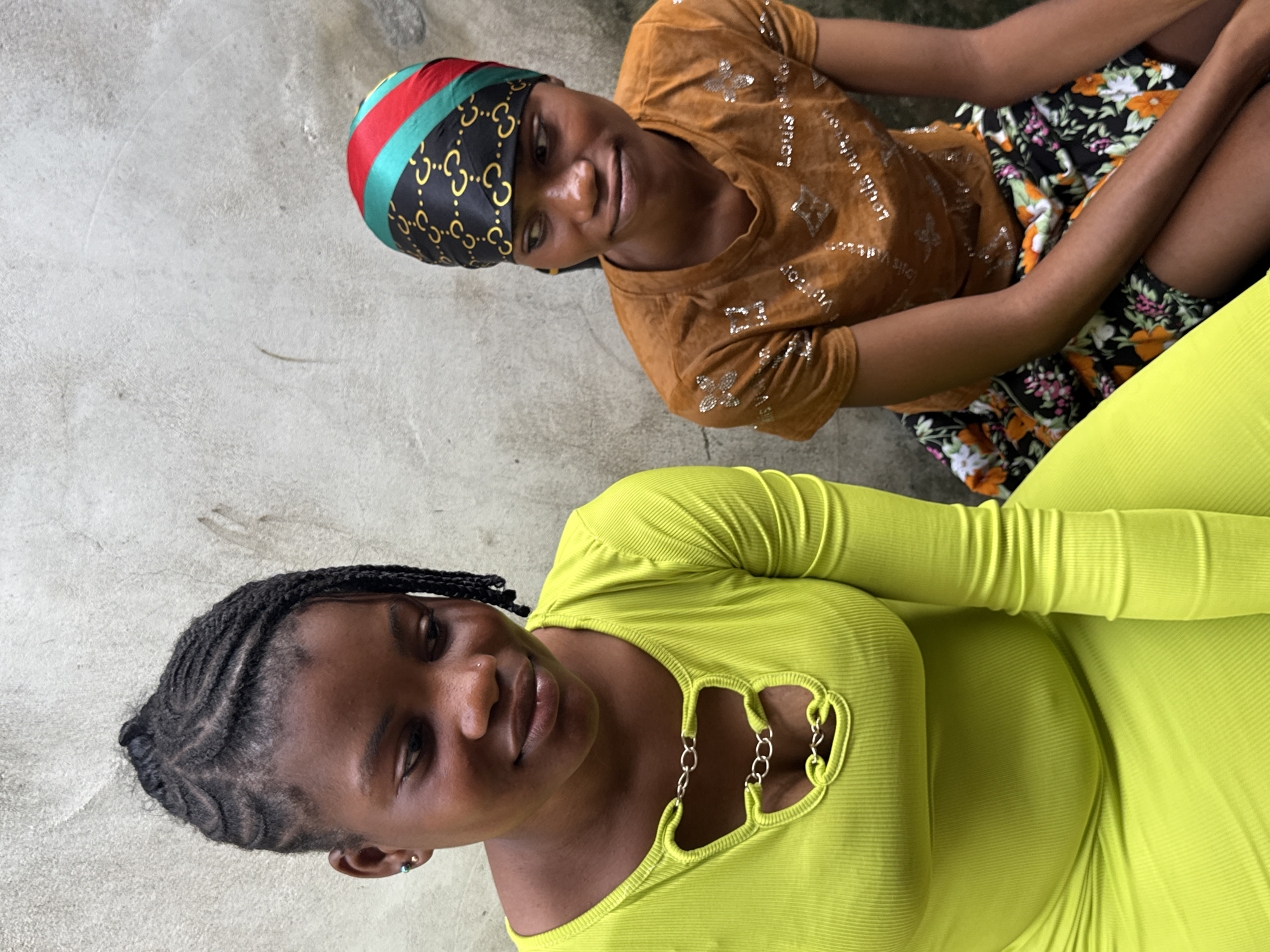 Two women are seated outdoors on a concrete surface. One woman wears a brown short-sleeved shirt, a floral skirt, and a colorful headscarf. The other woman wears a light green, form-fitting, long-sleeved dress with a decorative chain detail on the neckline. They appear to be posing for the camera. The setting is outdoors, possibly in Badagry, Nigeria.