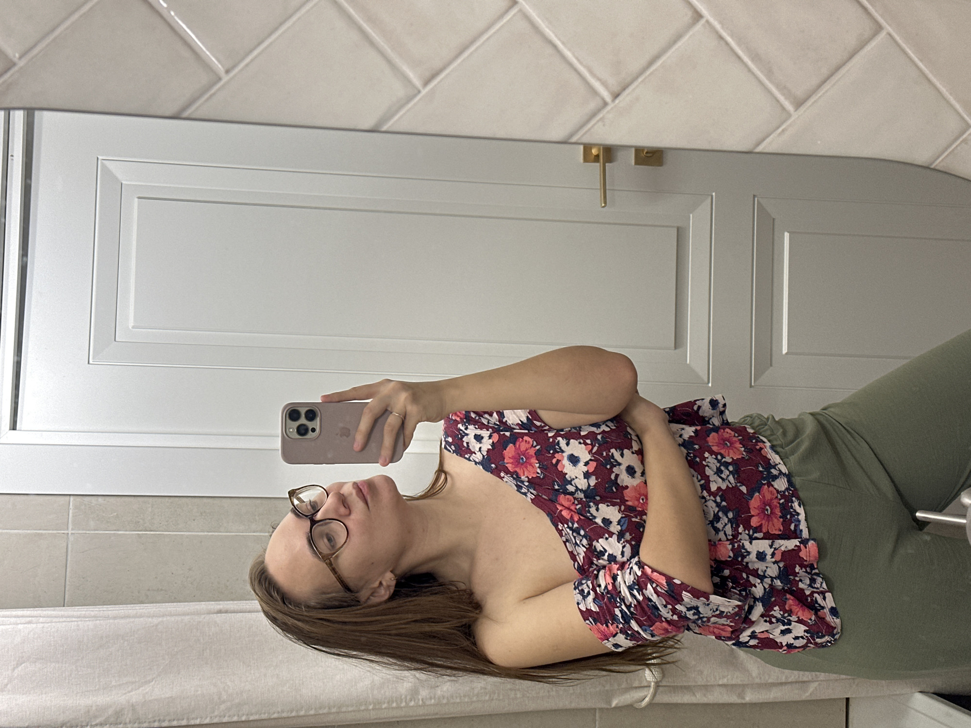 A woman with brown hair and glasses is taking a selfie in front of a mirror. She is wearing a floral shirt and green pants. The background shows a white door and herringbone tile. The location is not discernible as Kharnavy, Russia.