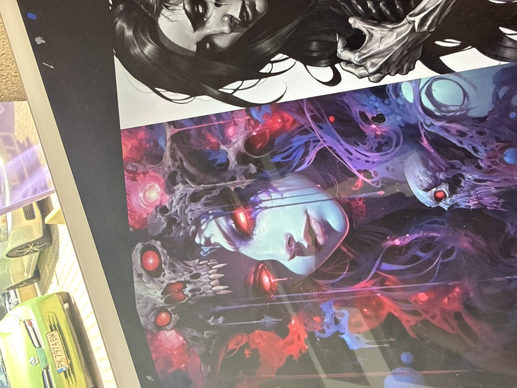 The image shows two pieces of artwork stacked on top of each other, possibly posters or prints, sitting on a surface that includes part of a toy car with a Bulgarian license plate "PK 7974 BH". The top artwork depicts a woman with dark hair, rendered in black and white. The artwork underneath is in full color and depicts a woman with pale skin and red eyes, surrounded by skeletal figures and swirling purples and reds. The license plate on the car indicates that this image was likely taken somewhere in Bulgaria; however, the location cannot be more precise than Bulgaria based on the available data.