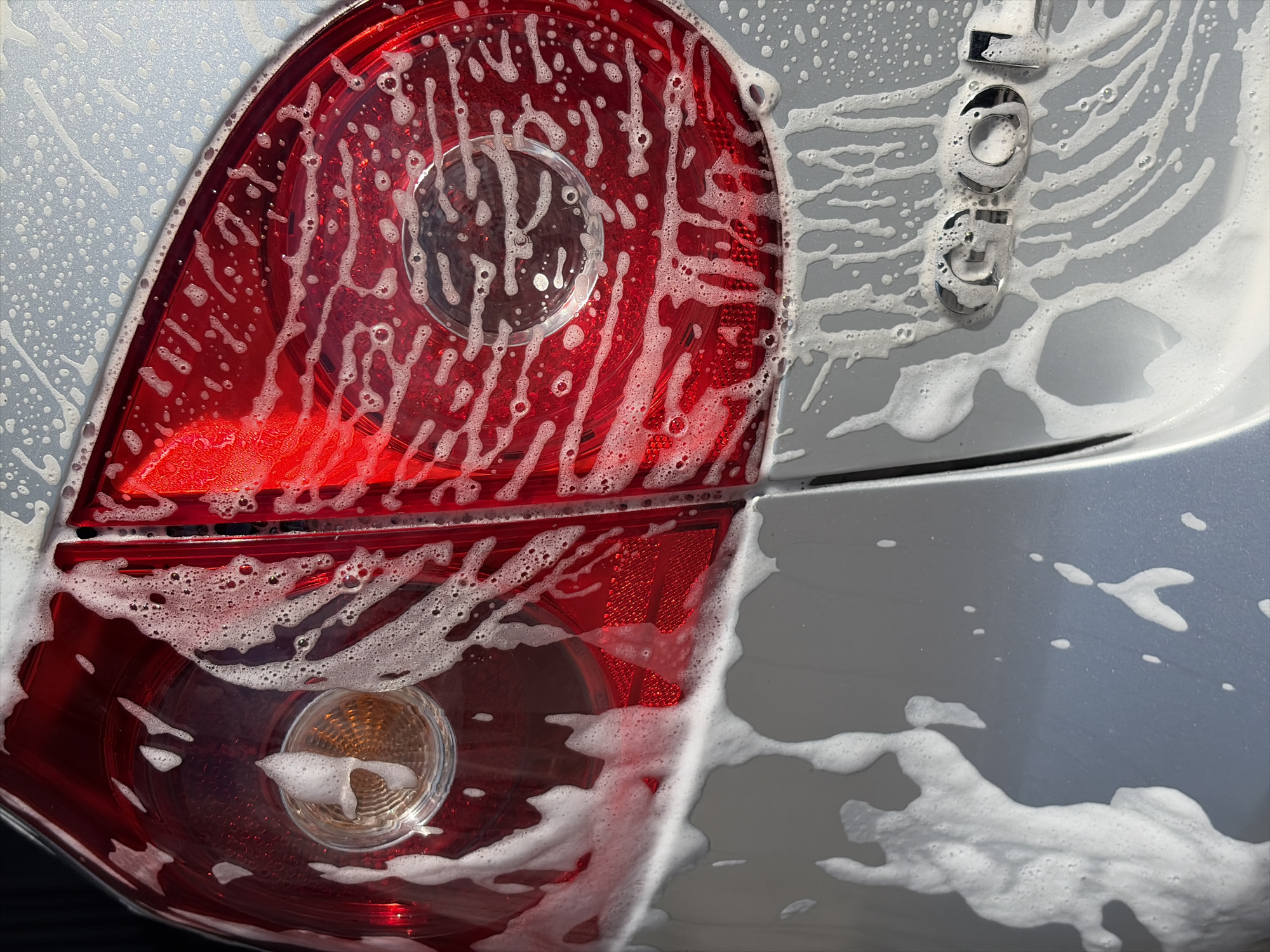 The image shows the rear passenger side of a silver car being washed. The car's taillight is visible on the left, covered in soap suds. The word "SOUL" is visible on the right, also covered in soap suds. The car appears to be in a car wash or being washed manually. The location is Unknown, Unknown.
