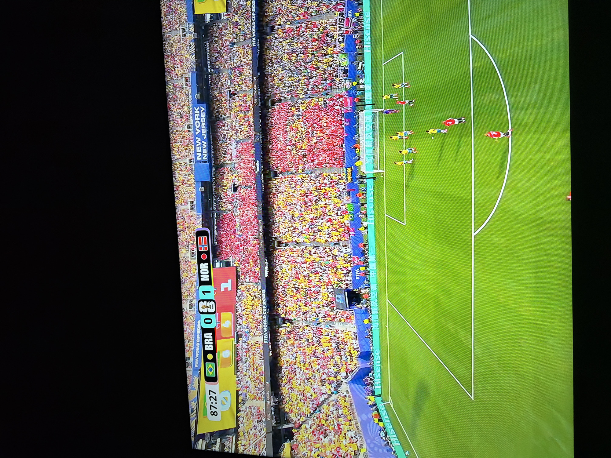 The image shows a soccer game in progress on a bright green field with white boundary lines. There are several players on the field, wearing yellow and red jerseys. The scoreboard at the top of the image indicates that the game is between Brazil (BRA) and Norway (NOR), with Brazil at 0 and Norway at 1. The time is 87:27. A large crowd is visible in the stands, with many spectators wearing yellow and red, suggesting strong support for one of the teams. Above the stands, a banner reads "NEW YORK NEW JERSEY". The image appears to be a capture of a television broadcast, as indicated by the display of the game's score and time. The scene is set outdoors, under natural lighting conditions. The specific location in Ognianovo, Bulgaria is not discernible from the image itself.
