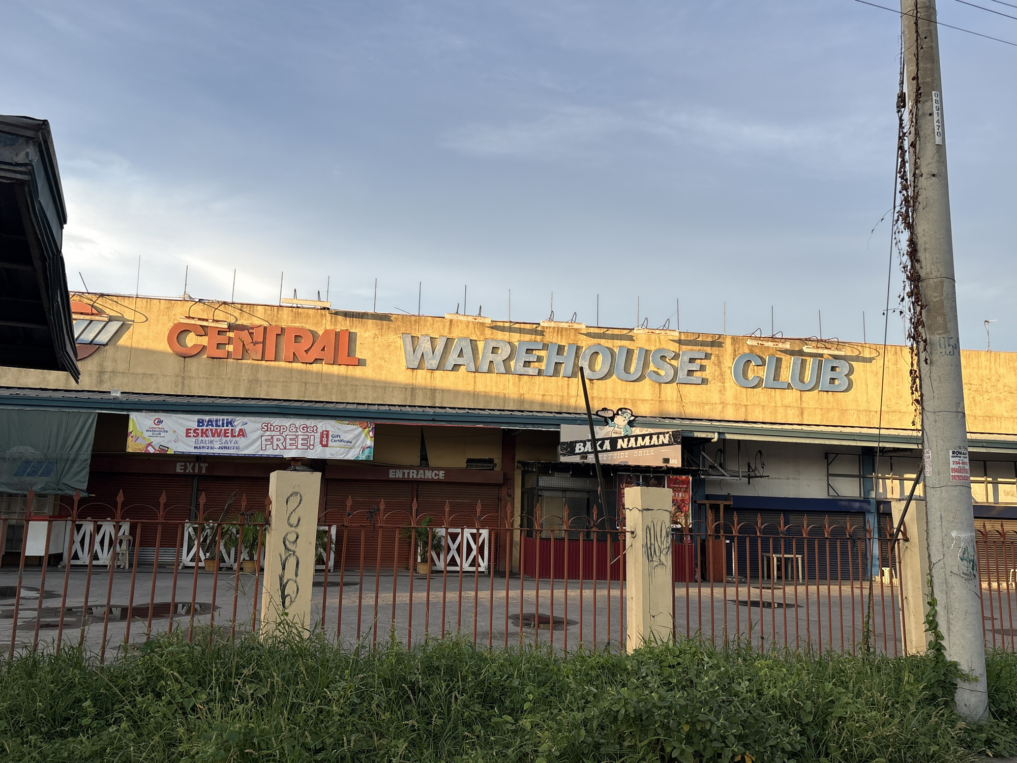 The image shows the facade of a building with the sign "CENTRAL WAREHOUSE CLUB" in large, orange and blue letters. The building is a commercial establishment, possibly a department store or a wholesale store. The exterior walls are painted yellow, and the roofline has several antennas. In front of the building, there is a red metal fence, and behind it, there are several rolled-up shutters, suggesting that the establishment might be closed or in a state of disrepair. A banner with text in Filipino and English is hanging above the entrance, advertising a "Balik Eskwela" (Back to School) event. There is also a sign that says "Baka Naman Streetside Grill". The scene is set against a backdrop of a cloudy sky, with the sun casting a warm glow on the building. The overall impression is one of a commercial area in the Philippines, possibly in Davao City given the mention of "Davao City, Philippines" in the prompt, though no explicit identifiers confirm this location. The scene is somewhat rundown, with some graffiti on a concrete pillar in the foreground.