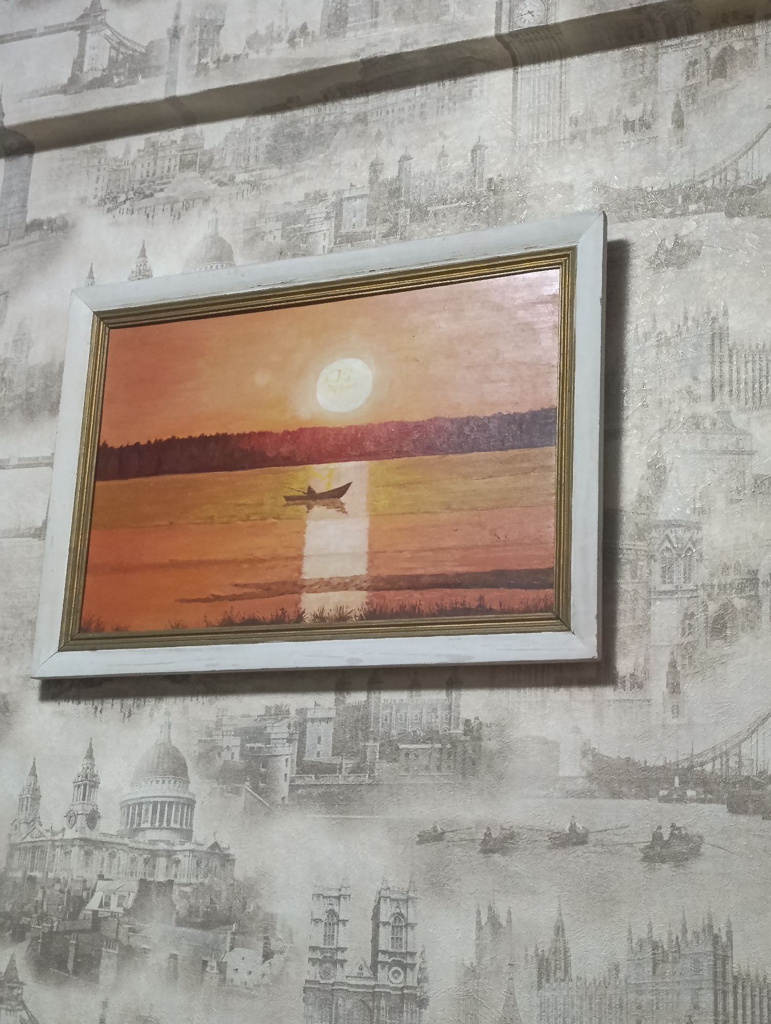The media file captures a framed painting hanging on a wallpapered wall in a room located in Krasnoyarsk, Russia.
The wallpaper features a detailed, monochromatic print of various iconic London landmarks and scenes. Recognizable structures include Big Ben (Elizabeth Tower) and the Houses of Parliament, St. Paul's Cathedral, and Tower Bridge. The pattern also shows multiple rowing boats with people on a river, likely the River Thames. The wallpaper has a textured finish, giving it a slightly aged or distressed look.
The framed painting, which has a white outer frame and a gold inner trim, depicts a serene waterscape at either sunrise or sunset. The sky and water are bathed in warm tones of orange, yellow, and red. A prominent, bright sun or moon is positioned in the upper right area of the sky, casting a strong, shimmering reflection across the water towards the foreground. Faint, irregular shapes, possibly resembling letters or symbols, are visible within the sun/moon. A small, dark-silhouetted boat with a single figure is situated on the water, slightly to the left of the sun's reflection. A dark band of trees or land forms the horizon line between the sky and the water, and subtle dark shapes, possibly reeds or distant land, appear at the very bottom edge of the painting.
The scene conveys a sense of tranquility and solitude. The time of day is clearly twilight, characterized by the dramatic lighting and warm color palette. No discernible text beyond the faint shapes within the sun/moon is visible in the painting.