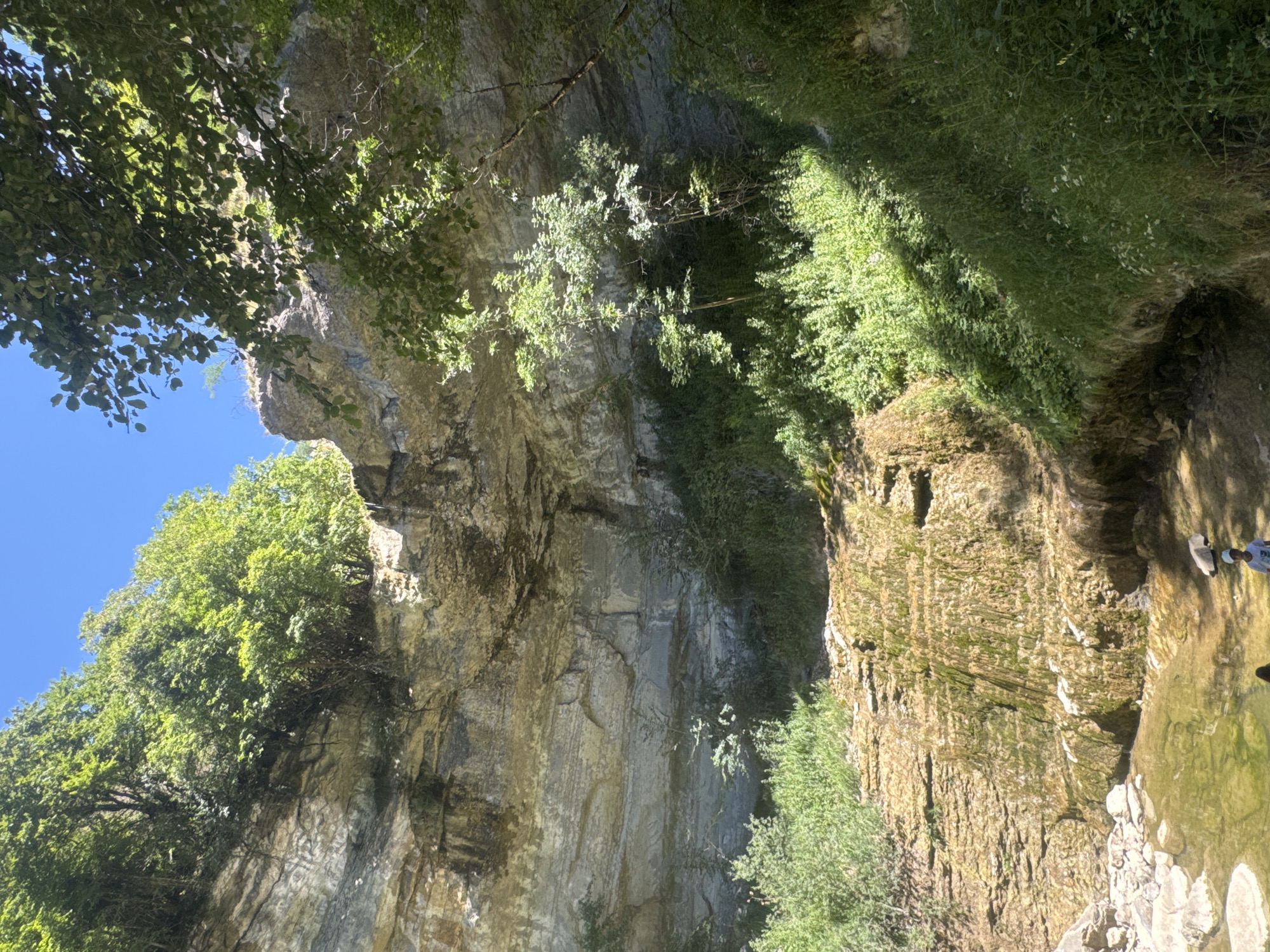 The image captures a natural scene featuring a small waterfall cascading down a rocky cliff face. Lush green vegetation covers the slopes and the top of the cliff, with sunlight filtering through the leaves of trees. At the base of the waterfall, a shallow pool of water is visible, with rocks scattered around its edge. Two individuals are present near the water's edge; one person is kneeling or sitting while the other stands. The scene suggests a natural, secluded area, likely a park or a natural reserve. This location is identified as the Cascade de Tacon, situated in Saint-Claude, France.