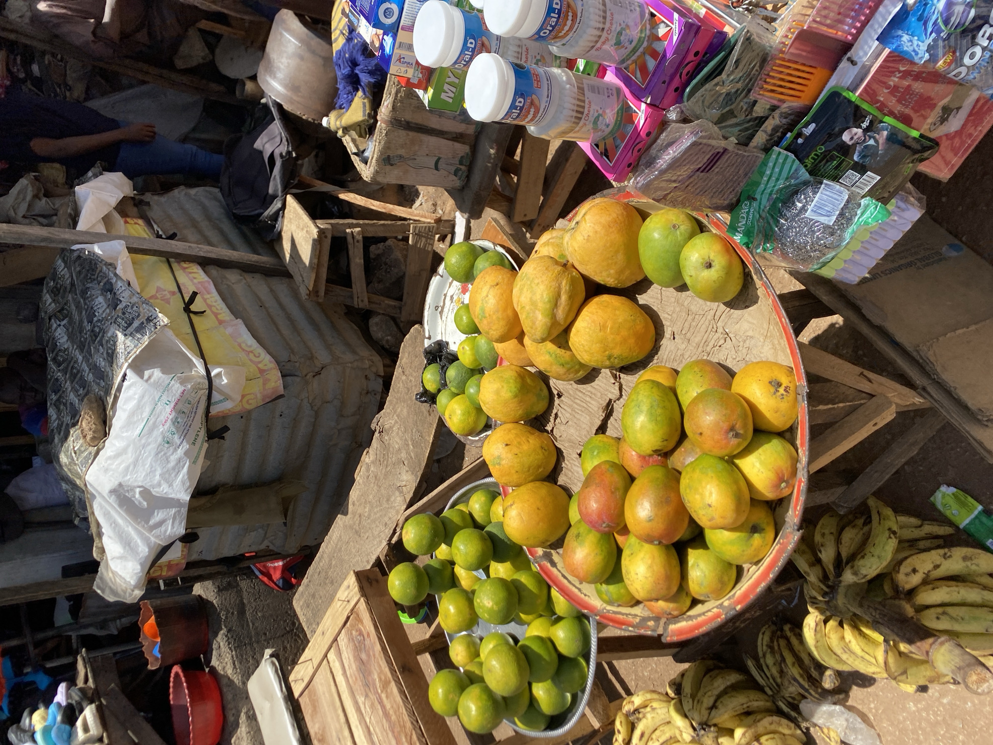 The image depicts a market scene in Bukuru, Nigeria. Assorted fruits, including mangoes, limes, and bananas, are displayed in bowls and trays. Various products like cotton swabs (branded "Oral-D") and packaged goods are arranged on wooden crates. Personal care items, cleaning supplies, and other goods are also visible. A person lies on the ground amidst the market's structures, which include corrugated metal sheets and wooden supports. The scene appears to be outdoors, lit by natural sunlight.