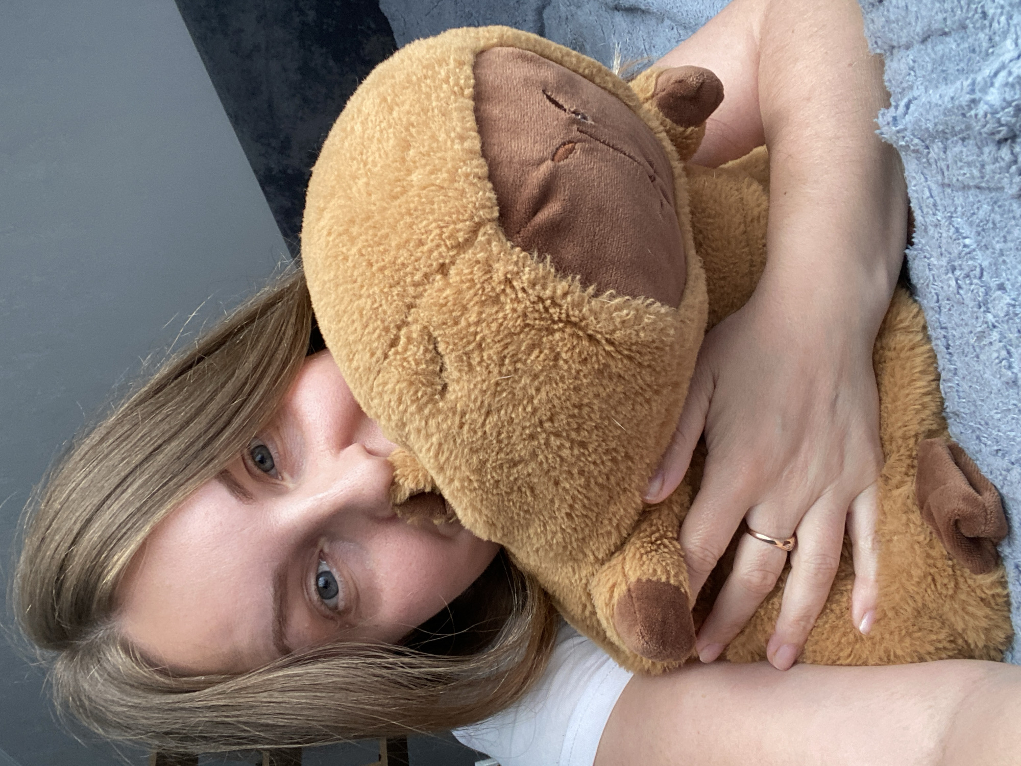 A light-skinned woman with blonde hair and blue eyes is holding a brown stuffed animal. She is wearing a white shirt and a gold ring on her finger. A blue blanket and a grey wall are visible in the background. The location is not discernible from the image.