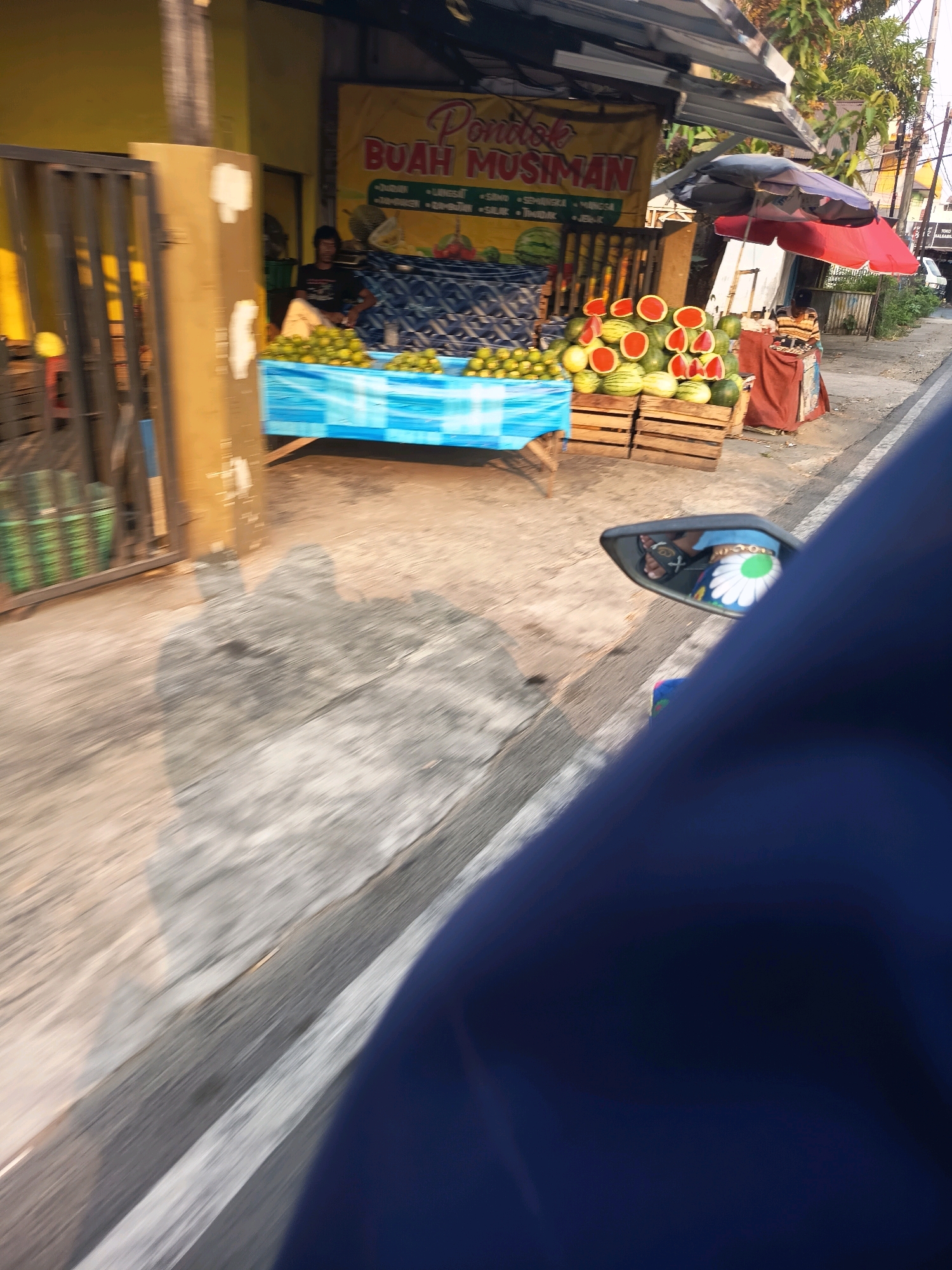 In Banjarmasin, Indonesia, a roadside fruit stand is captured in motion, likely from a moving vehicle. The stand displays an assortment of fruits, including whole watermelons and halved watermelons showing their red interiors, piled in wooden crates and on a blue tarp. Oranges are also visible in green crates. A person wearing a striped shirt sits behind the fruit display under a red and black umbrella. A banner above the stand reads "Pondok Buah Musiman" and lists various fruits. The scene is characterized by the motion blur typical of a moving camera, with the road, sidewalk, and surrounding buildings creating a dynamic background. A side-view mirror reflects a person's legs adorned with a bracelet and a floral-patterned garment.