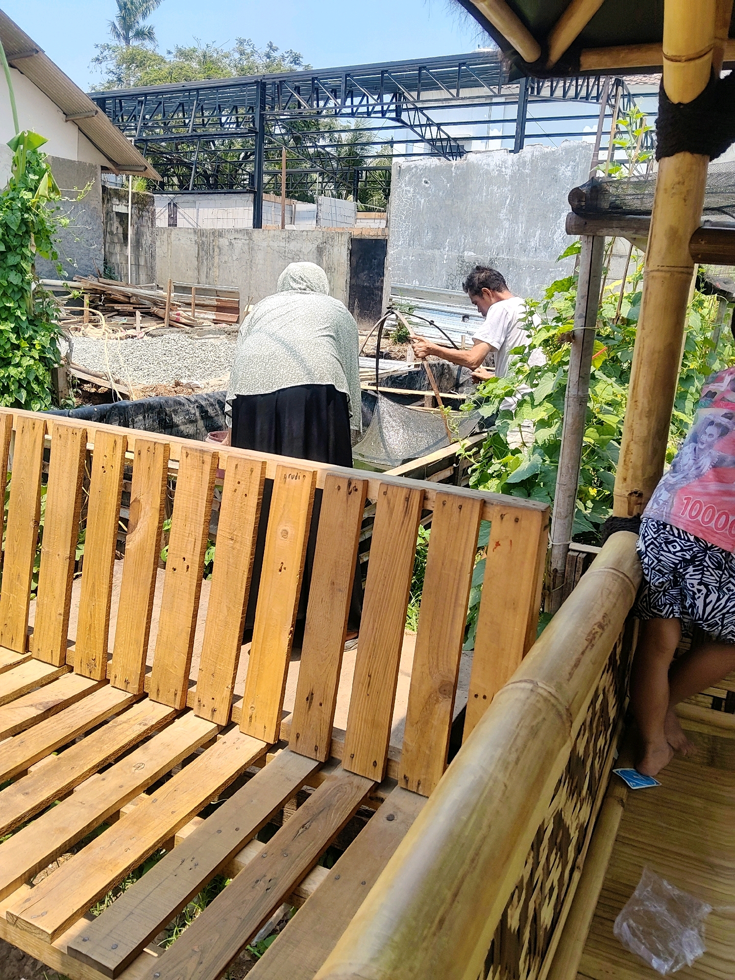 In Cibatu, Indonesia, a construction site is depicted under a clear sky. The foreground features a rustic wooden bench made of pallets. Beyond the bench, a person wearing a headscarf and a long skirt is seen from behind, tending to a pond. To their right, a man in a white t-shirt is engaged in some activity near the pond, holding a tool. In the background, a building structure is under construction, with exposed metal framing and concrete walls. Green foliage and trees surround the area, indicating an outdoor setting.