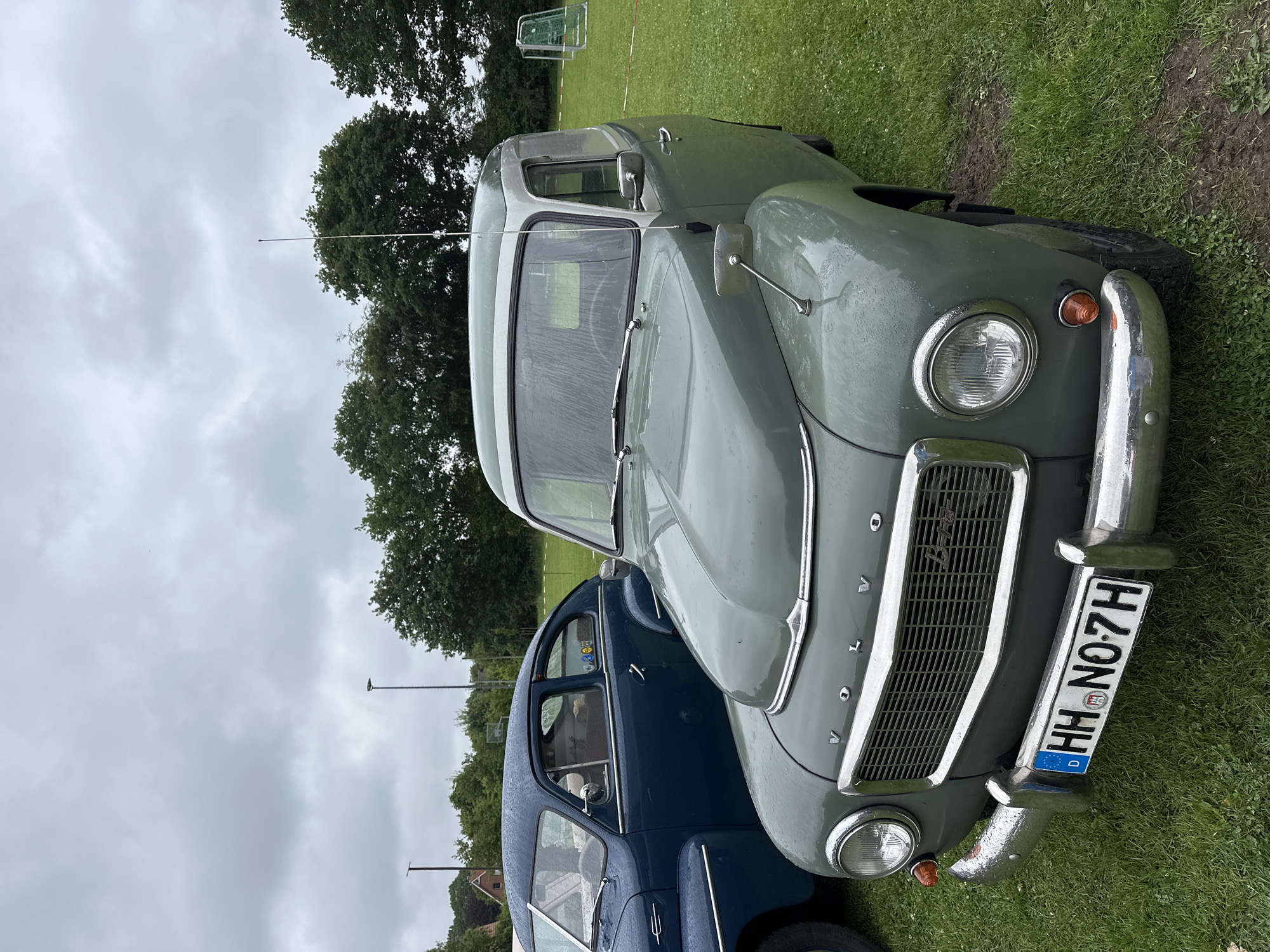 The image depicts two classic Volvo automobiles parked on a wet, green grass field under an overcast, cloudy sky in Deinste, Germany.
In the foreground, slightly angled towards the viewer from the right, is a two-tone grey-green Volvo PV445/210 Duett panel van or estate, covered in visible raindrops. Its front features a prominent chrome grille with "VOLVO" lettering above and a "B18" badge on the lower right section of the grille. Circular headlights with chrome bezels are positioned on either side of the grille, with amber turn signal lamps mounted below them on the bumper supports. The front bumper is chrome, showing some minor wear and reflections. A German license plate, "HH N0 7H," is centrally mounted on the front bumper, indicating the car is registered in Hamburg (HH) and featuring the Hamburg coat of arms. A long, slender antenna extends upwards from the front passenger side of the vehicle's lighter grey roof. A chrome side mirror is mounted on the driver's side front fender. The wet green grass beneath the vehicle shows some darker, muddy patches, particularly near the front wheel.
Behind and to the left of the prominent grey-green Volvo, the front-left section of a dark blue classic Volvo sedan (likely a PV544 or similar model) is partially visible. It also shows numerous raindrops on its surface and chrome trim around its windows. Small yellow and black stickers are affixed to its rear-left side window.
The background consists of a line of dense, dark green deciduous trees stretching across the mid-ground. Above the trees, the sky is uniformly grey with heavy cloud cover, suggesting a damp or rainy day. Several tall, dark streetlights or utility poles are visible above the tree line. In the far distance on the left, a building with a reddish-brown roof can be faintly seen. To the far right, two white soccer goalposts are visible, standing on the grass field, with a segment of white and red striped barrier tape on the ground nearby. No people are visible in the scene.